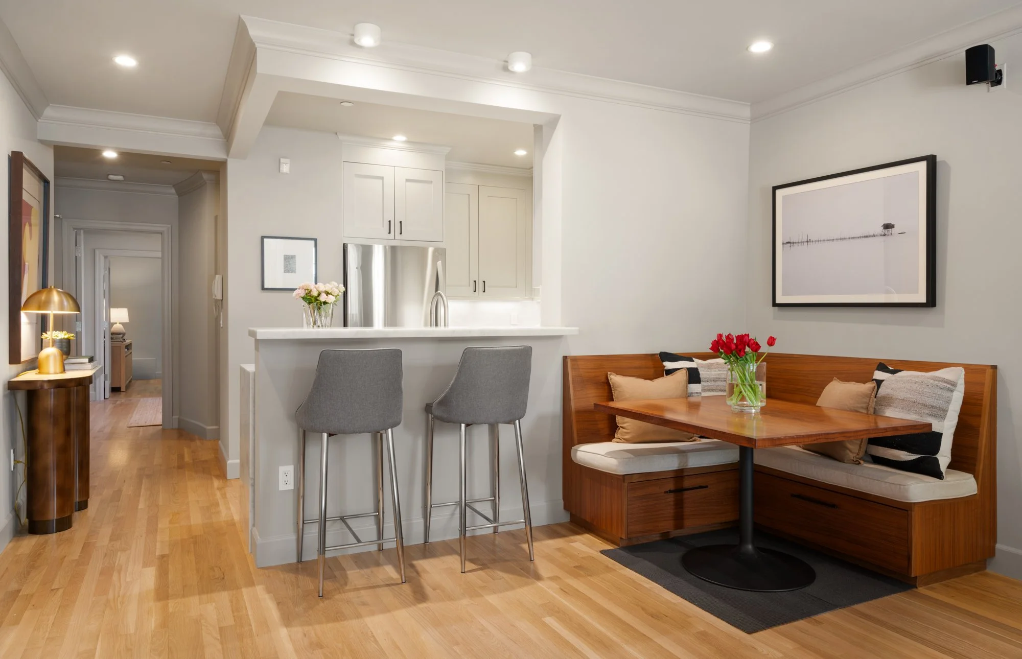 A cozy living room with a wooden dining nook, a small table with cushions and a vase of red tulips, a white kitchen with cabinetry, a stainless steel refrigerator, two gray barstools at a white counter, framed wall art, and a hallway leading to other