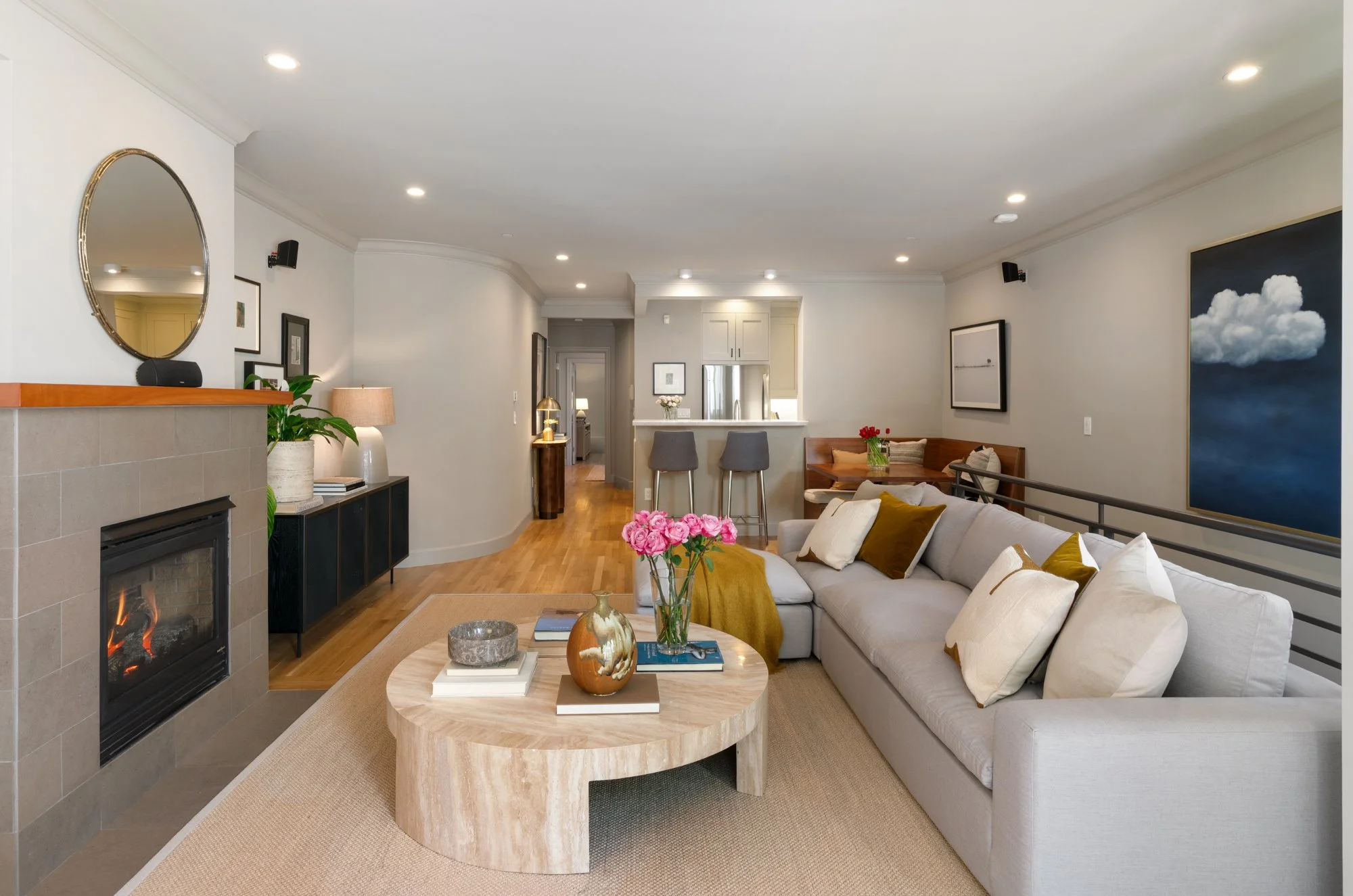 Living room with a white sofa decorated with yellow and white pillows, a round beige coffee table with books, vases, and decorative items, a gas fireplace with a beige tiled surround, a black media console with potted plant and lamps, artwork on the 