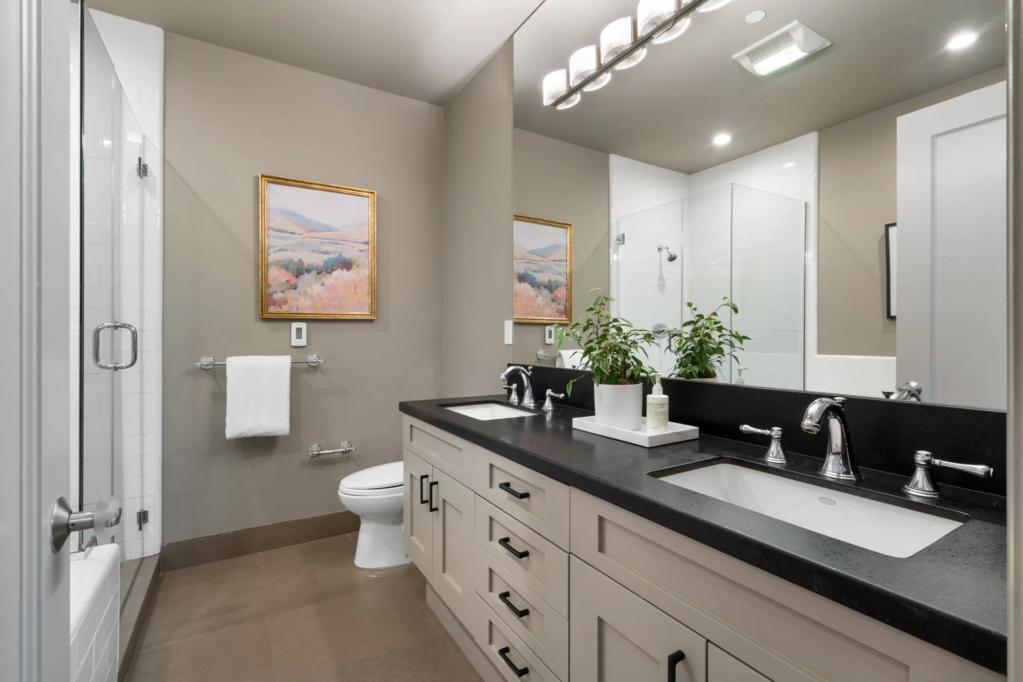 Modern bathroom with a double vanity featuring a black countertop, white cabinets, and two sinks. There is a mirror above the vanity, a plant on a tray, and transparent containers with cotton balls. The bathroom includes a glass-enclosed shower, a to
