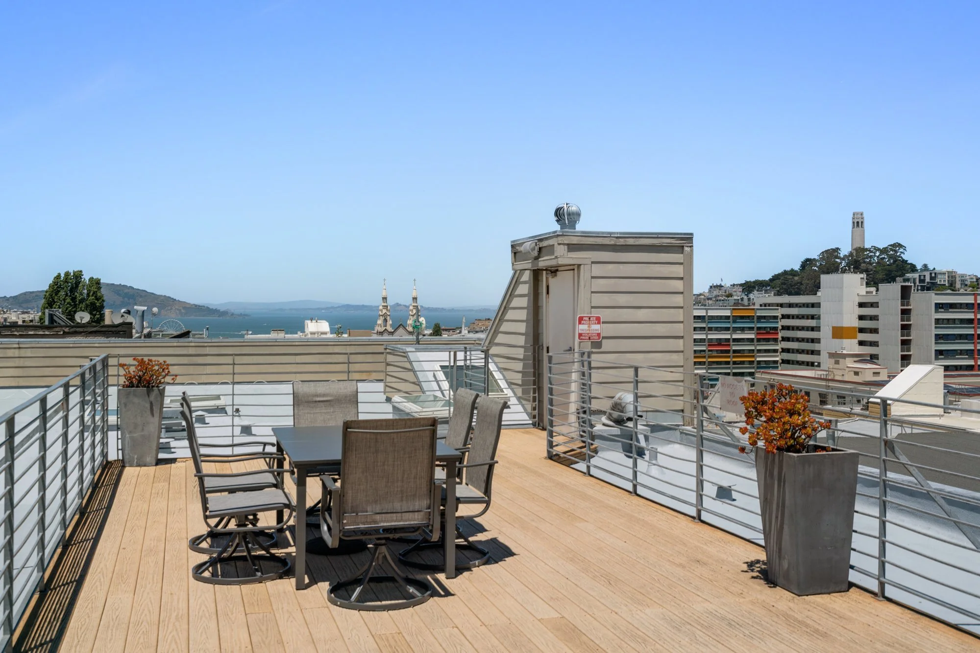 Rooftop terrace with dining tables and chairs, potted plants, city skyline with church steeples, a tower, and a body of water in the background under a clear blue sky.