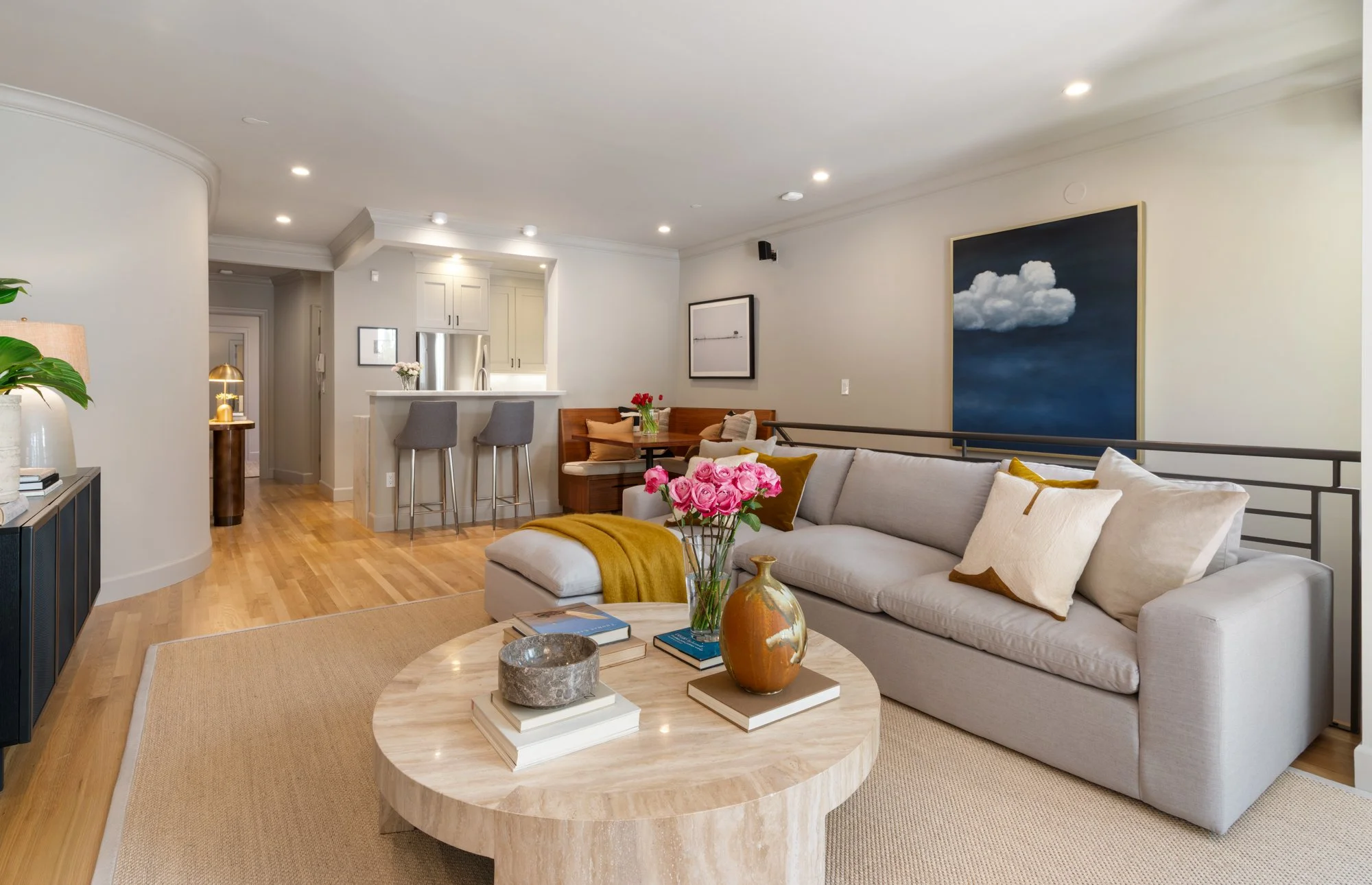 Living room with light-colored sofa, pink roses on a round coffee table, and artwork on the walls, adjacent to an open kitchen area with bar stools.