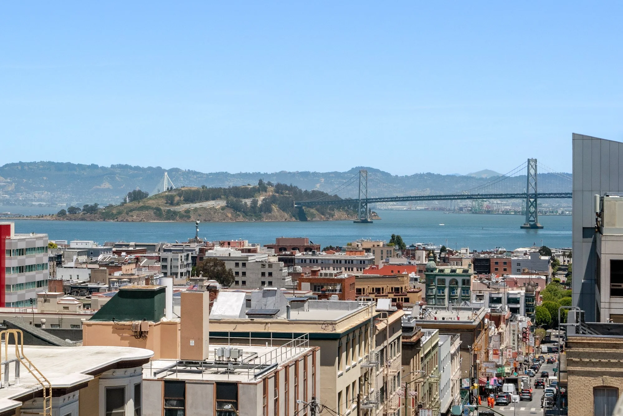 Cityscape with a bridge over water, hills in the background, and city buildings in the foreground under a clear blue sky.