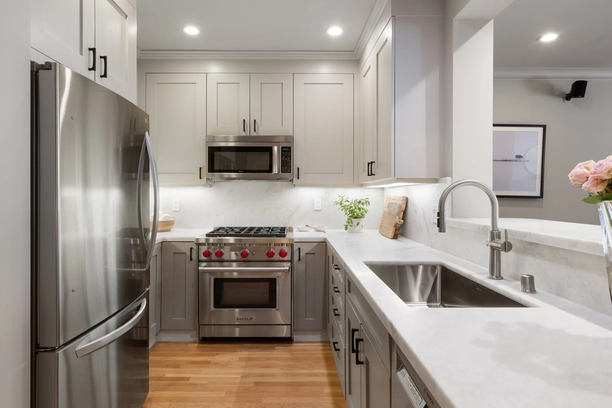 Modern kitchen with white cabinets, stainless steel appliances including a refrigerator, microwave, and oven, a white marble countertop with a sink, a small potted plant, and pink flowers in a vase.