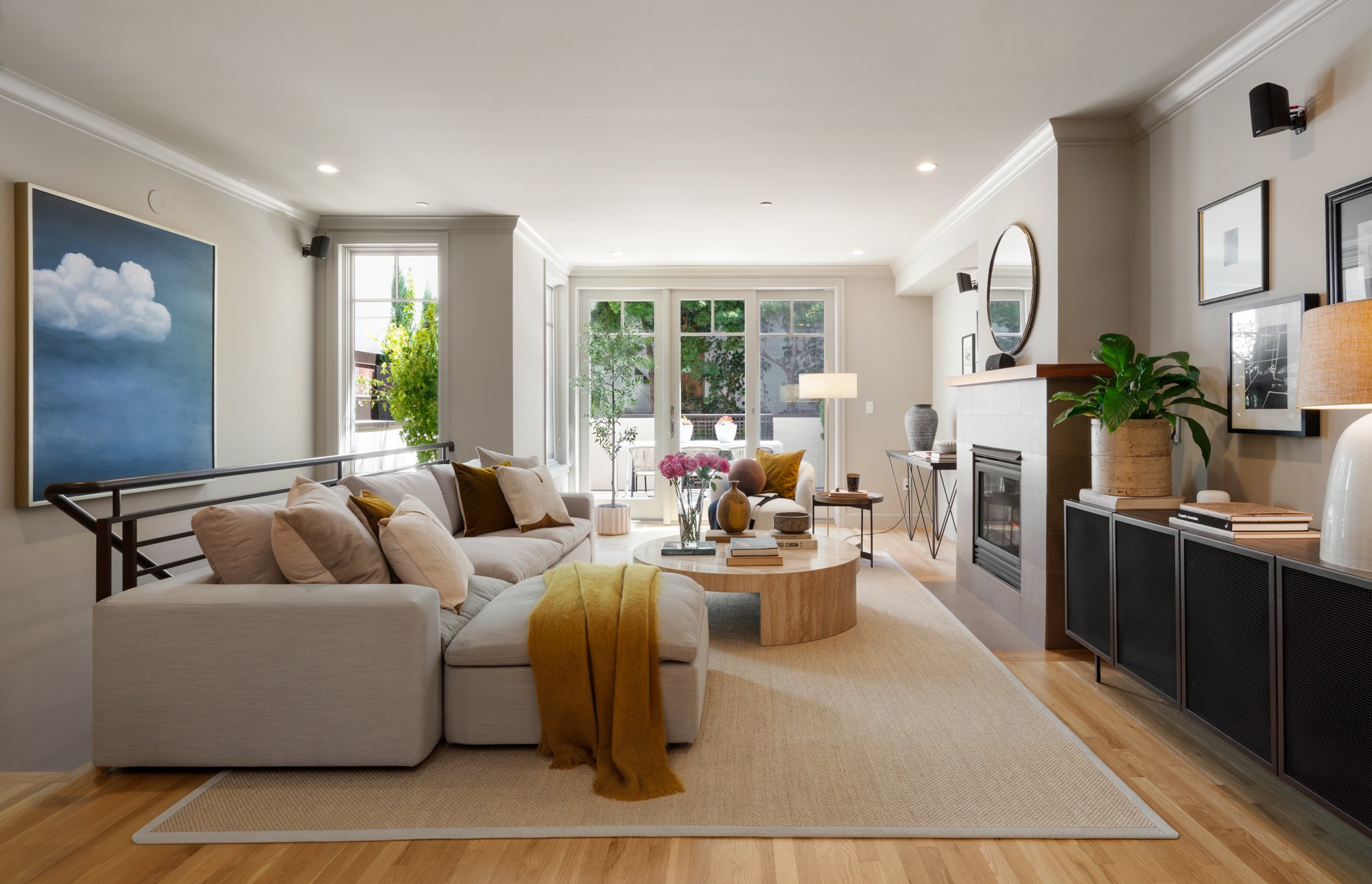 Bright living room with white walls, large windows, beige sofa with pillows, coffee table with decorative items, and wall art.