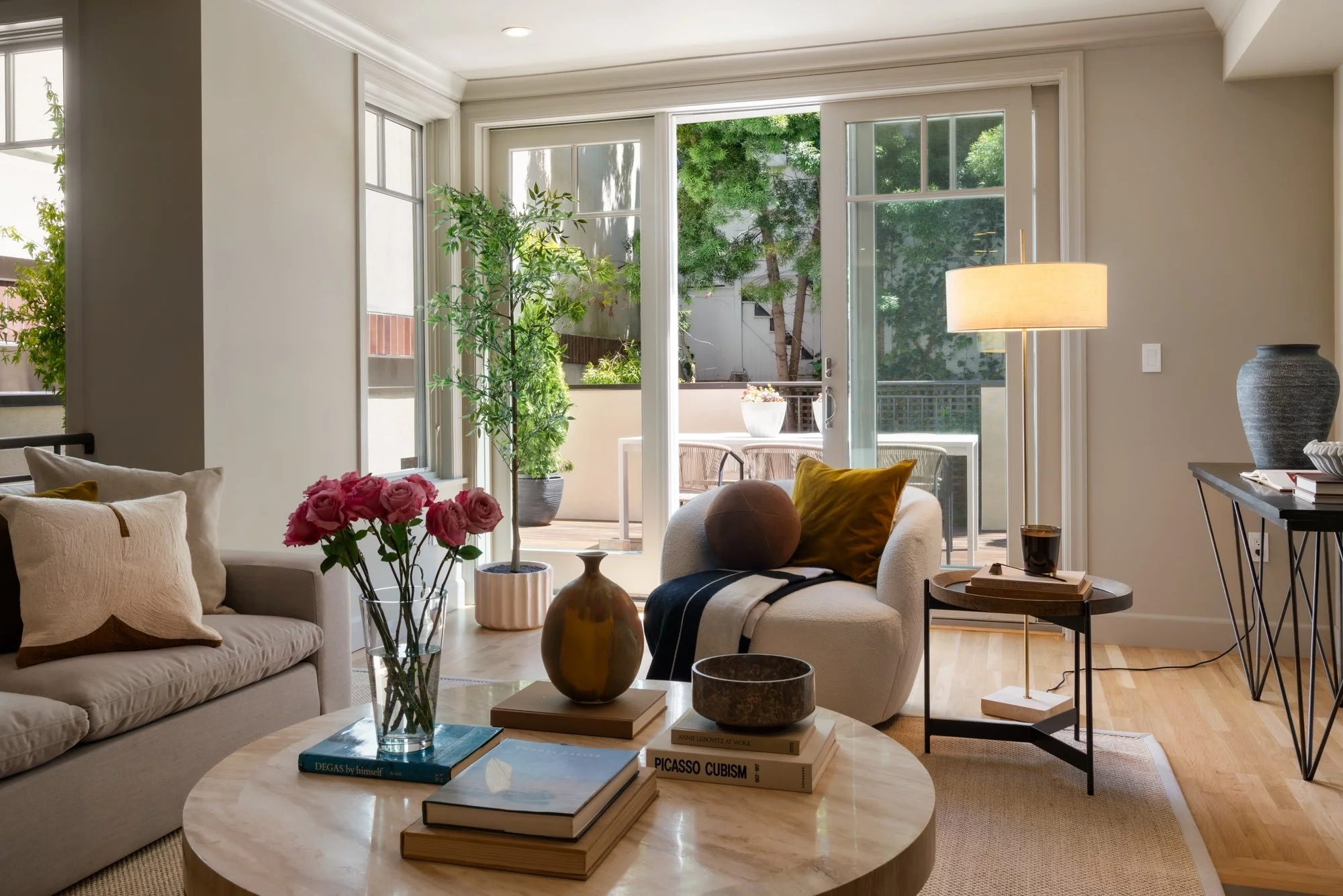Living room with beige couches, a glass vase with pink roses, various decorative books, and a windowed door leading to a balcony with greenery outside.