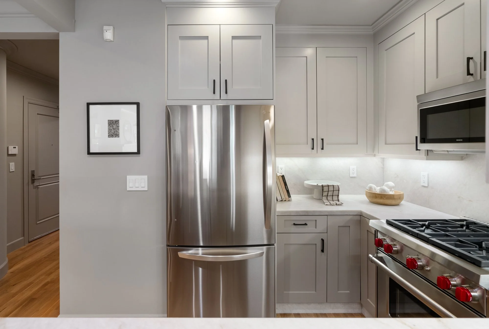 Modern kitchen with gray cabinets, stainless steel refrigerator, microwave, and stove, with wooden flooring and minimalist decor including books, a bowl with cotton balls, and a striped dish towel.