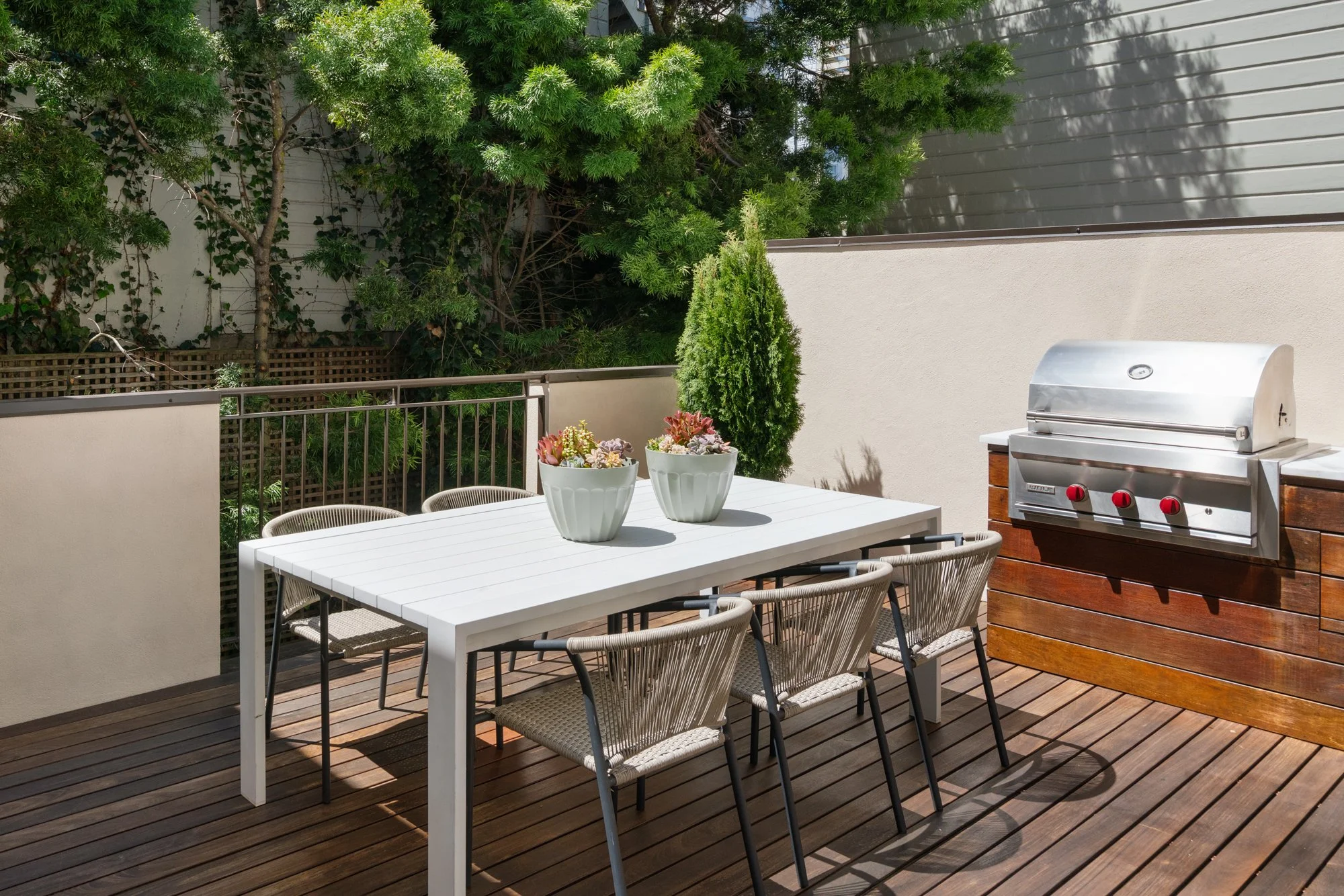A modern outdoor patio with a white dining table, six beige chairs, potted plants, a stainless steel BBQ grill, and lush green trees and shrubs in the background.