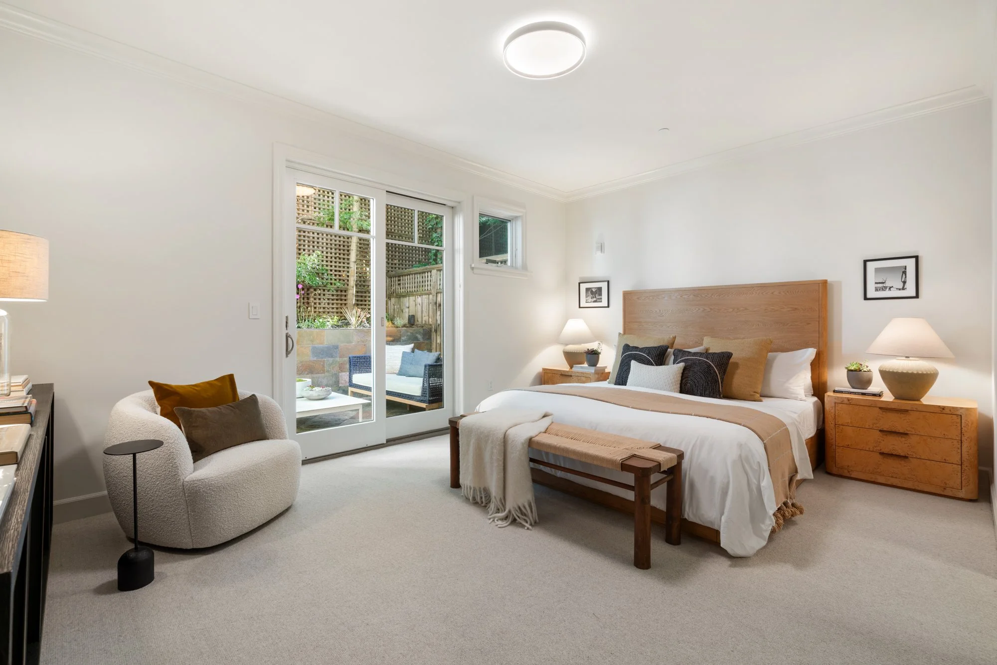 Modern bedroom with sliding glass door opening to an outdoor patio, featuring a bed with beige and black pillows, two wooden nightstands with lamps, a cream-colored armchair with pillows, a small side table, and framed black-and-white photos on the w