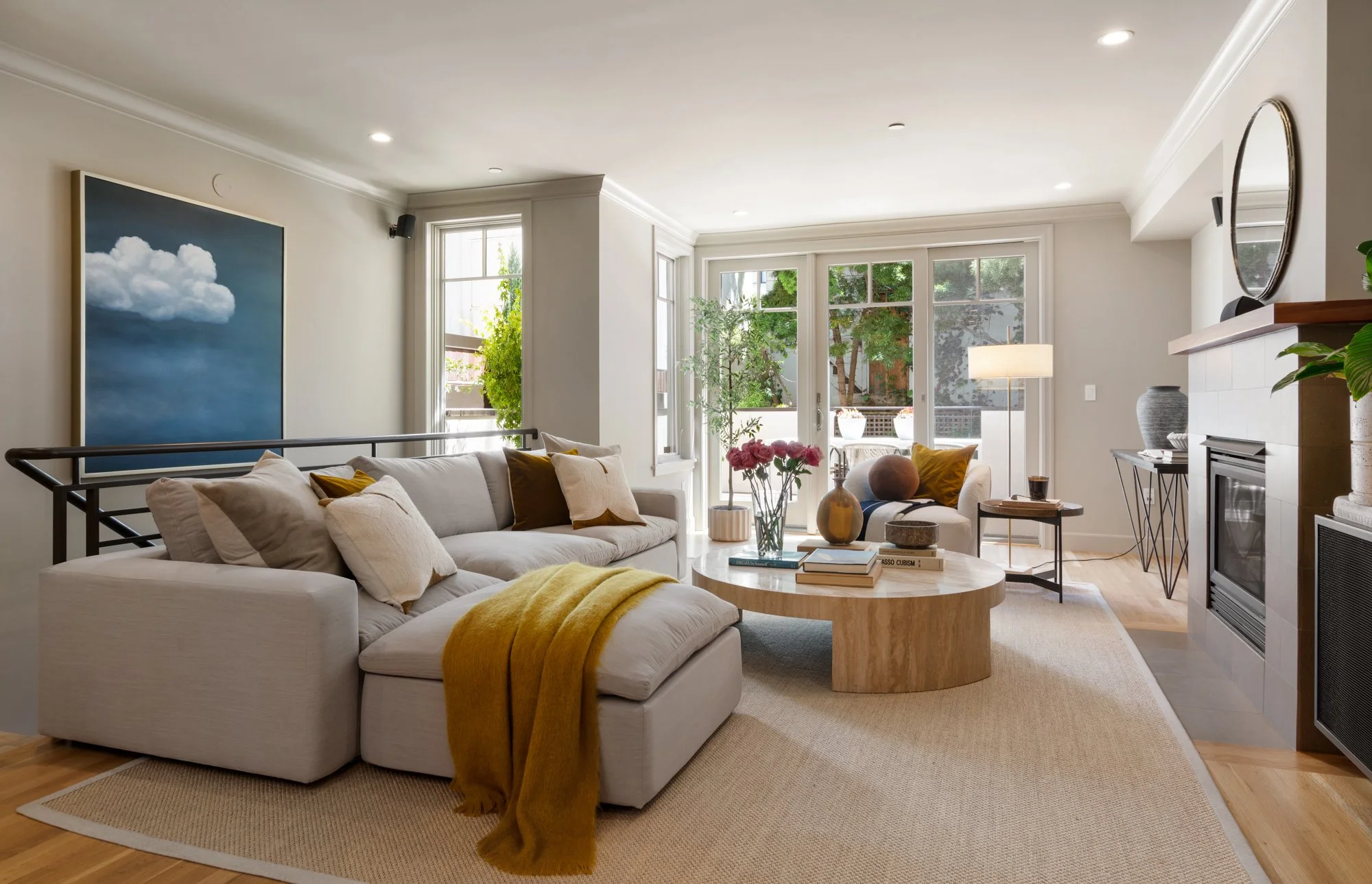 A bright living room with a large white sofa decorated with various pillows and a yellow blanket, a wooden coffee table with books and flower vases, and windows with views of greenery outside, complemented by indoor plants and modern decor.
