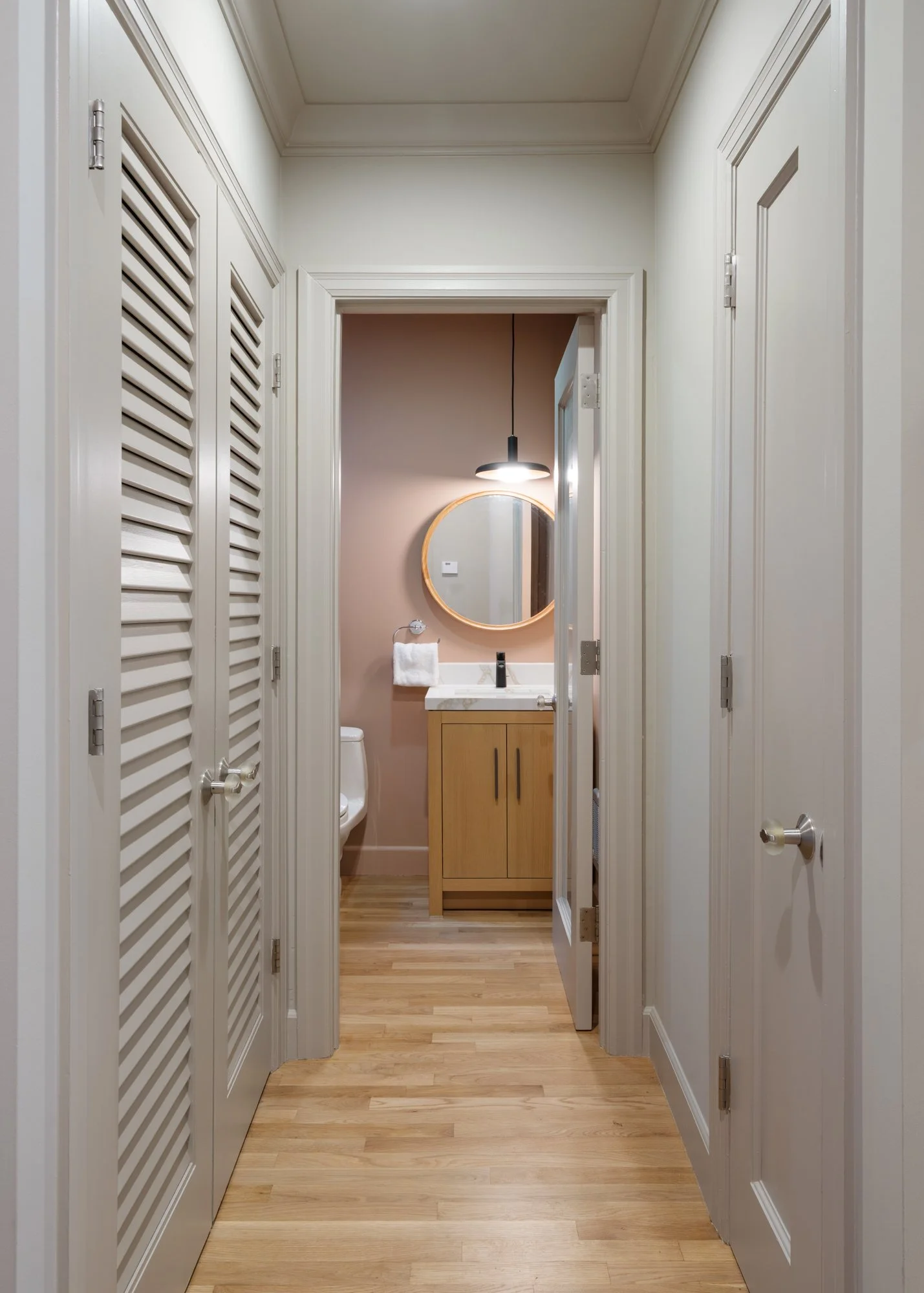 A clean, narrow hallway with wooden flooring, white walls, and white louvered closet doors on both sides leading to a small bathroom. The bathroom features a circular mirror, a black pendant light, a small wooden cabinet, a white sink, and a towel ha