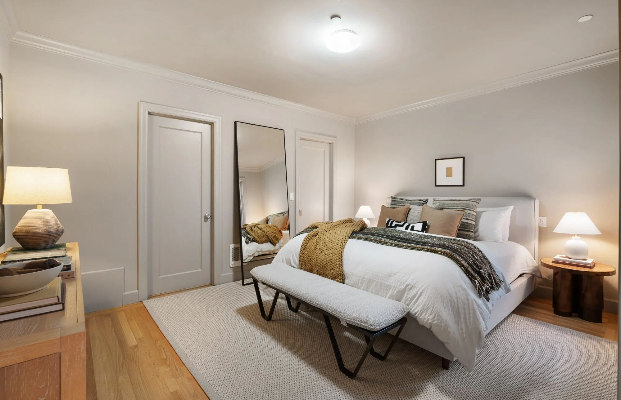 A bedroom with a white bed, beige, gray, and brown pillows, a yellow and brown striped blanket, a black and white throw, and a bench at the foot of the bed. There are wooden nightstands with lamps on each side, a tall mirror leaning against the wall 