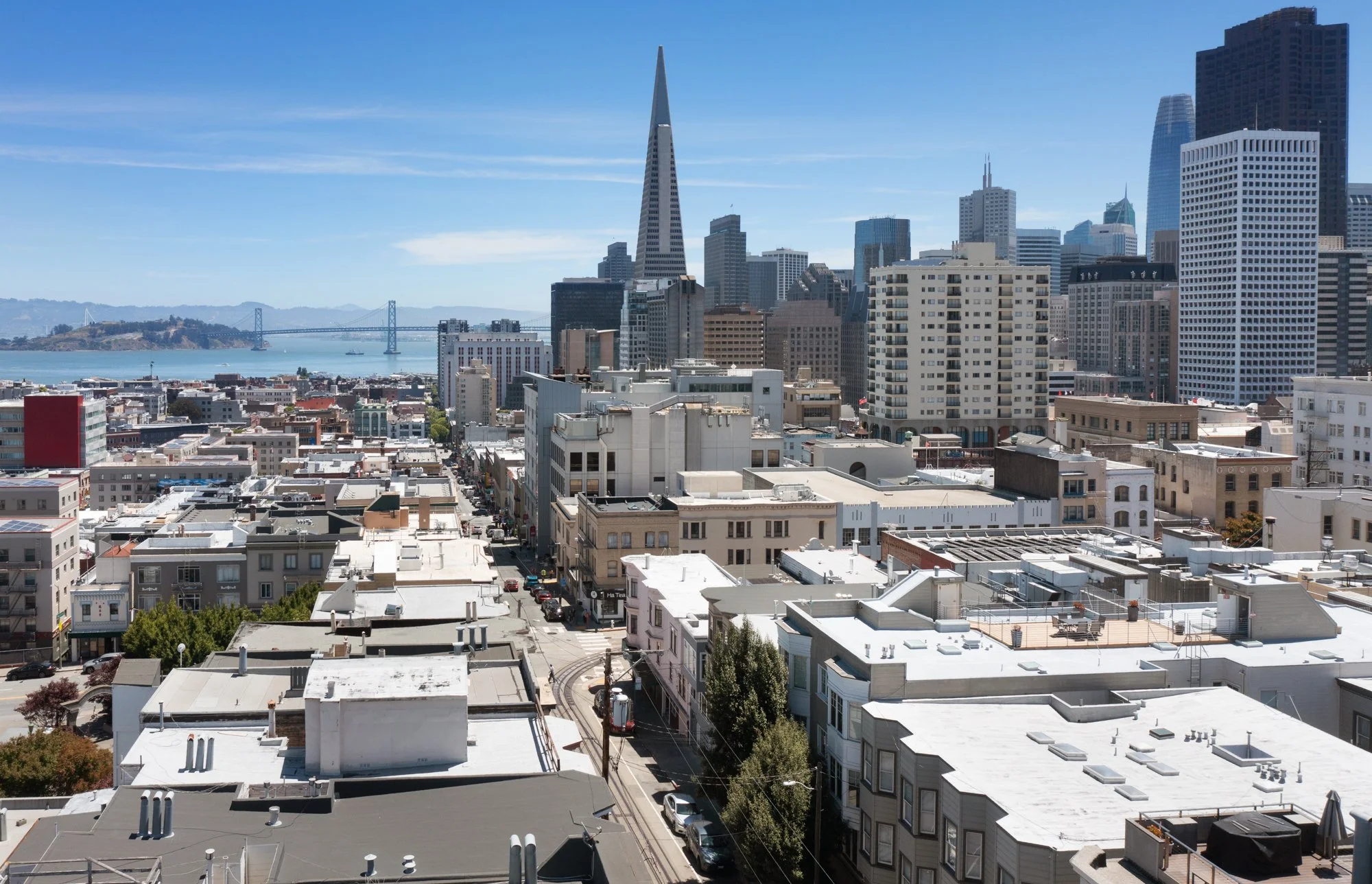 A cityscape view of downtown San Francisco with skyscrapers, the San Francisco Bay, and the Bay Bridge under a clear blue sky.
