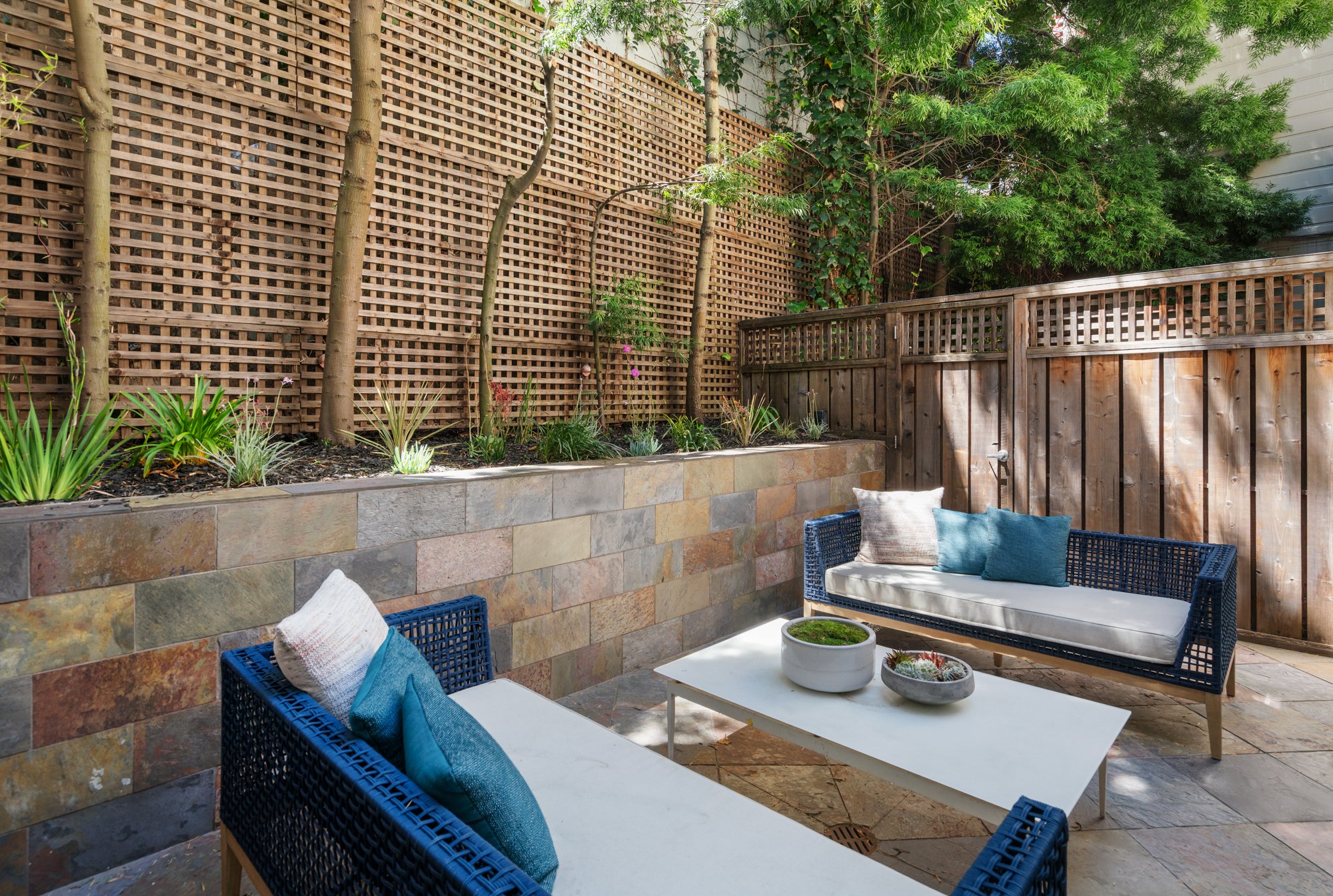 An outdoor patio area with woven blue chairs, white cushions, and pillows. There is a small white table with potted plants, a natural stone wall, wooden fencing, and a lush green tree in the background.