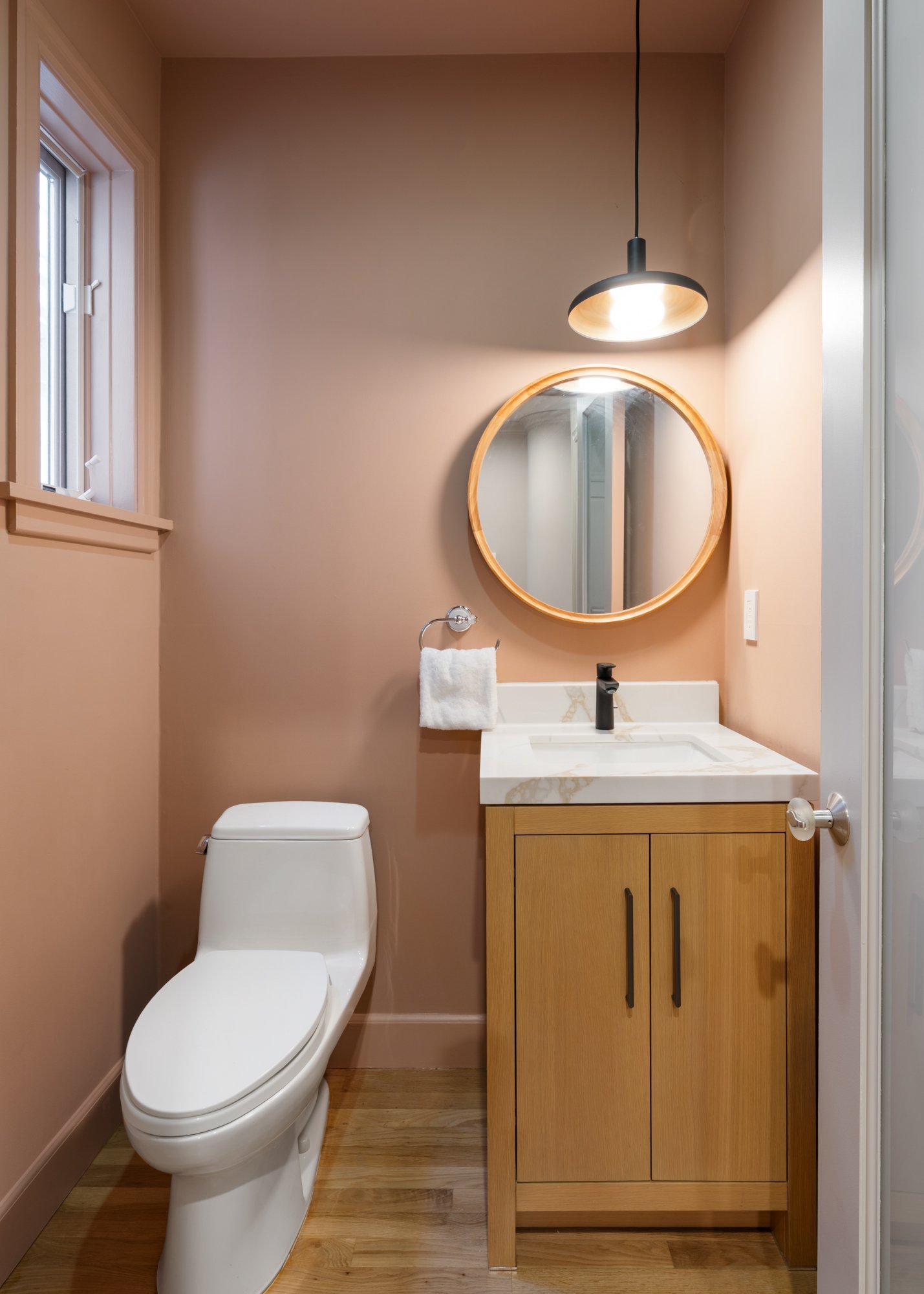 A modern bathroom with a white toilet, a wooden vanity with a white marble countertop, a round mirror above the sink, a black faucet, a towel ring with a white towel, a window on the left, and a pendant light hanging from the ceiling.
