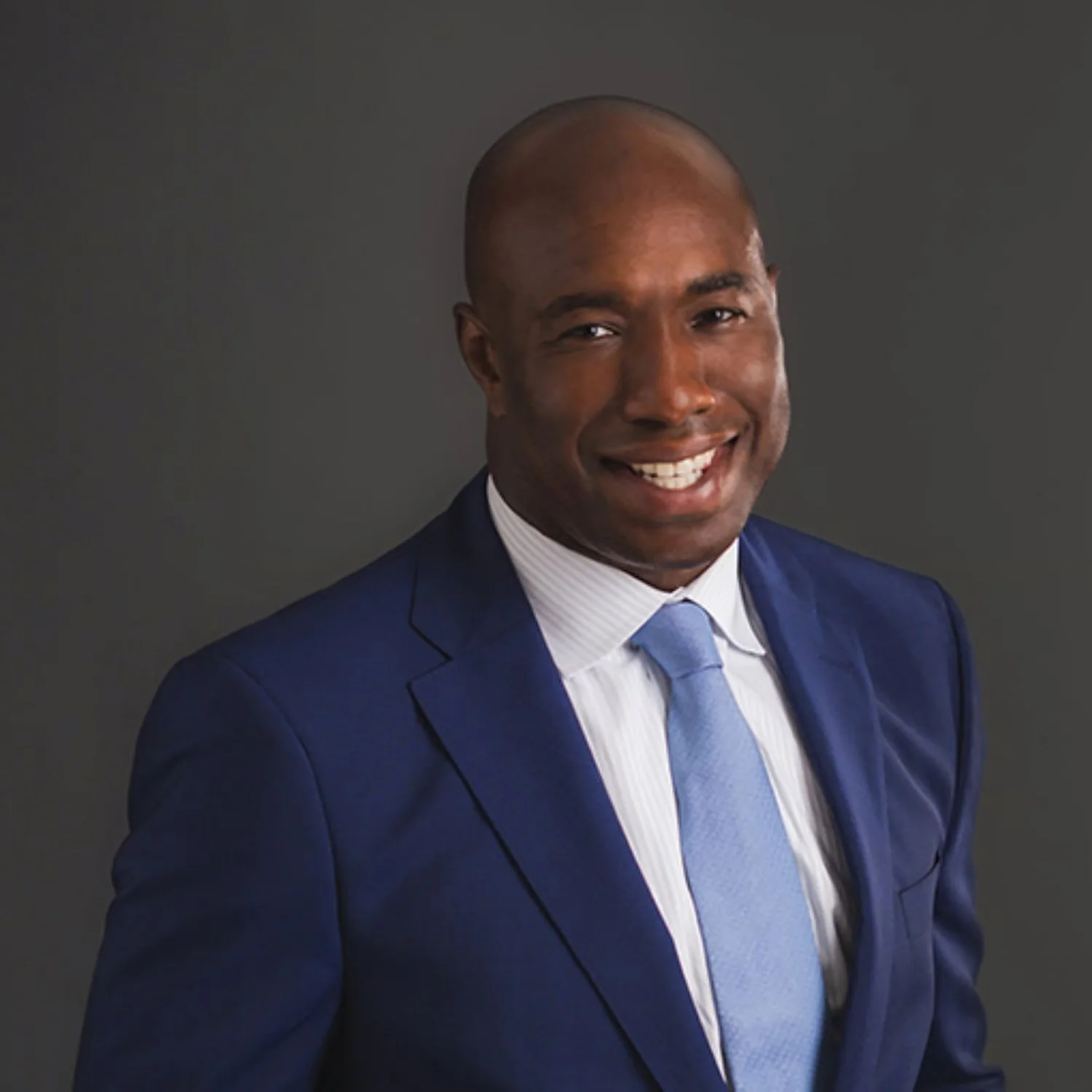 A smiling man in a blue suit, white shirt, and blue tie against a gray background.