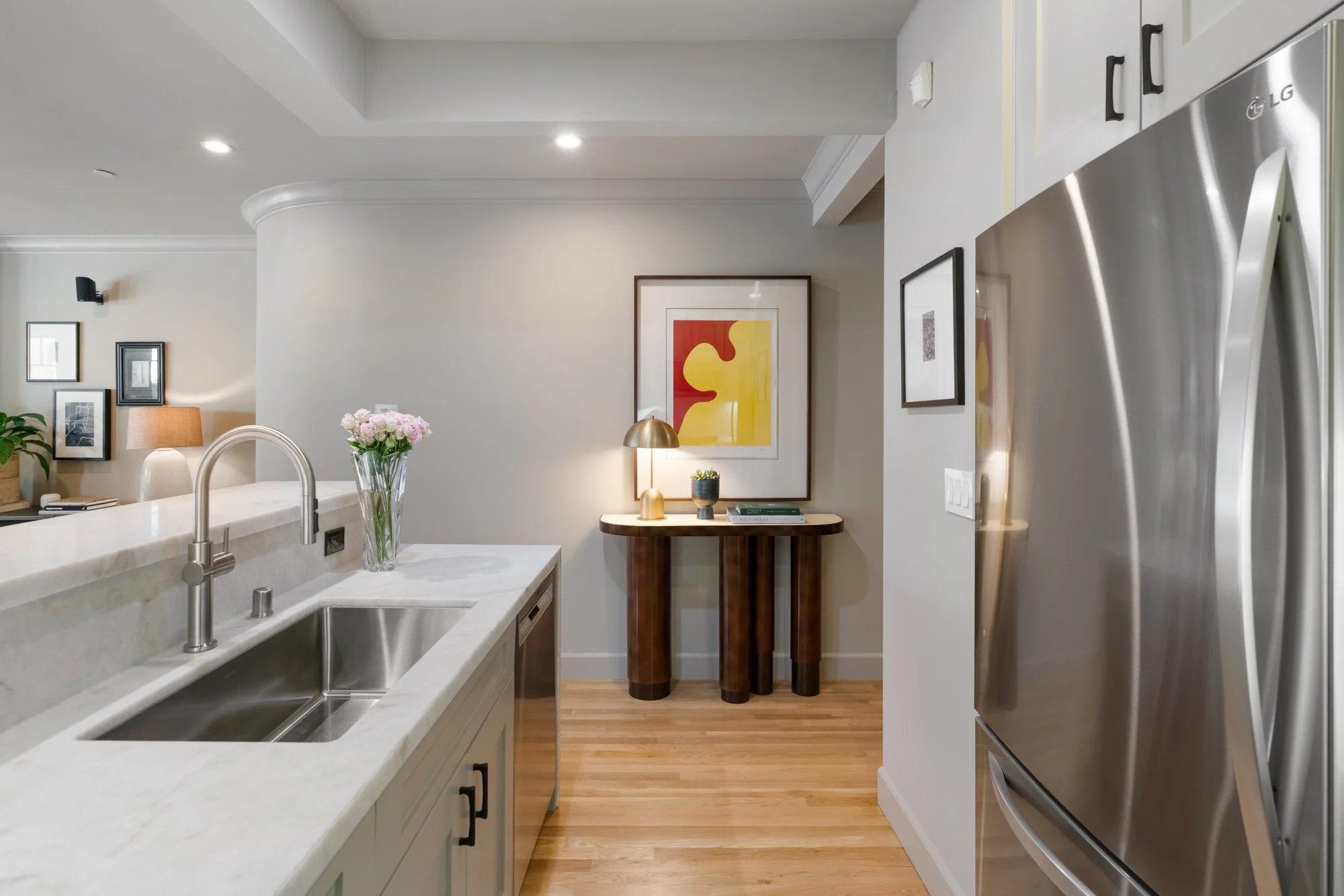 Modern kitchen with white cabinets, a stainless steel refrigerator, and a marble countertop with a sink. There are framed artworks and a table with a vase of pink flowers and a small lamp in the background.