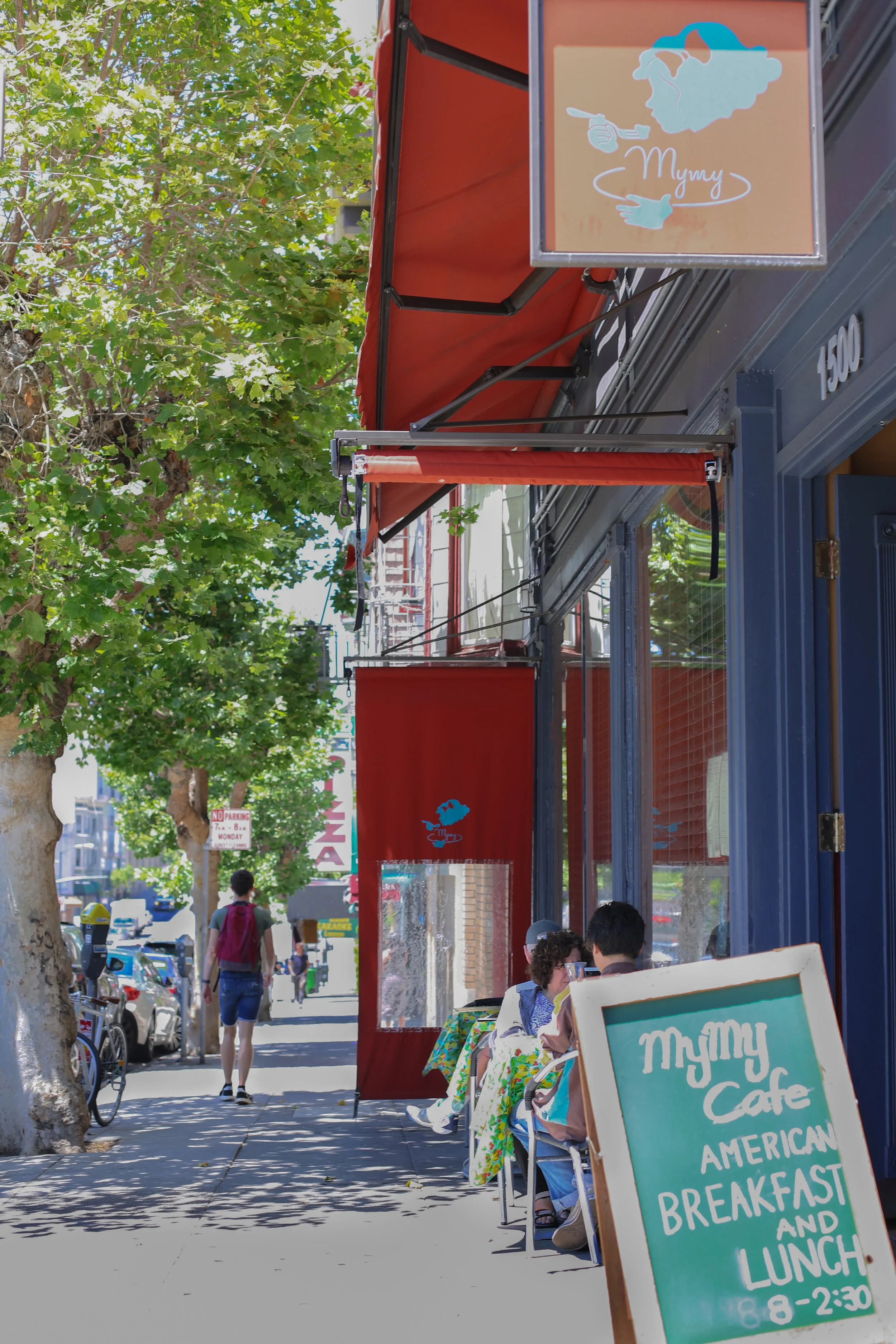 Street scene with people sitting at a cafe with bright red awnings, trees lining the sidewalk, and pedestrians walking by. Signs indicate a cafe serving American breakfast and lunch from 8 am to 2:30 pm.