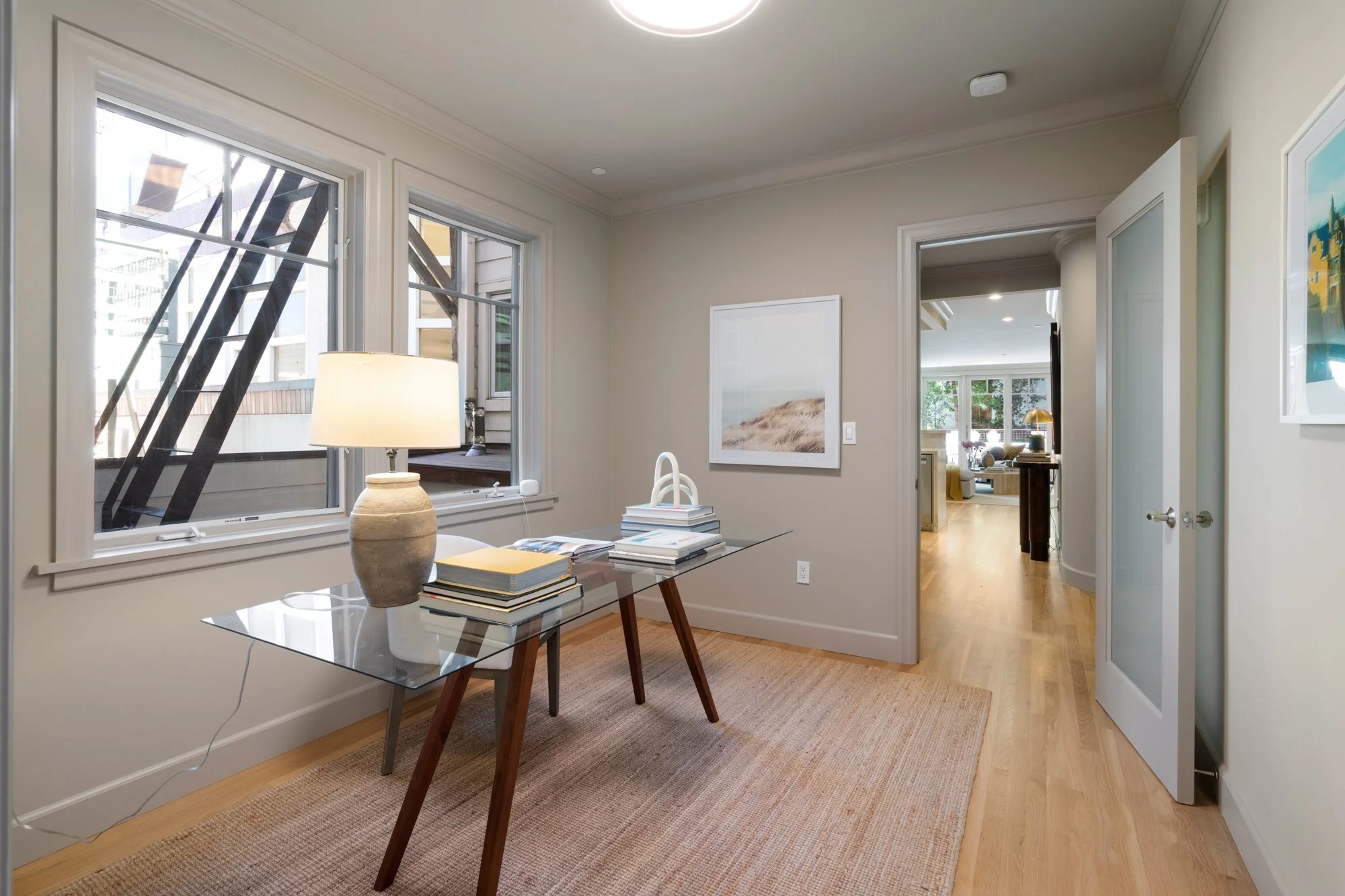 A bright, modern dining area with two large windows showing exterior buildings and fire escape stairs. There is a glass dining table with stacked books, a beige ceramic lamp, and a lamp on various tables in the adjacent living room. The room has ligh