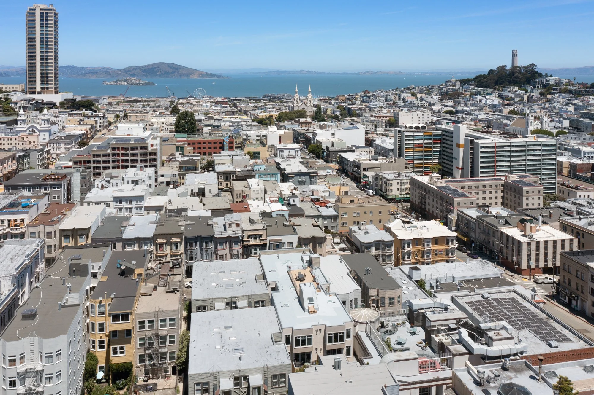 Aerial view of a city with a mix of residential and commercial buildings, a body of water in the background, and hills on the horizon.