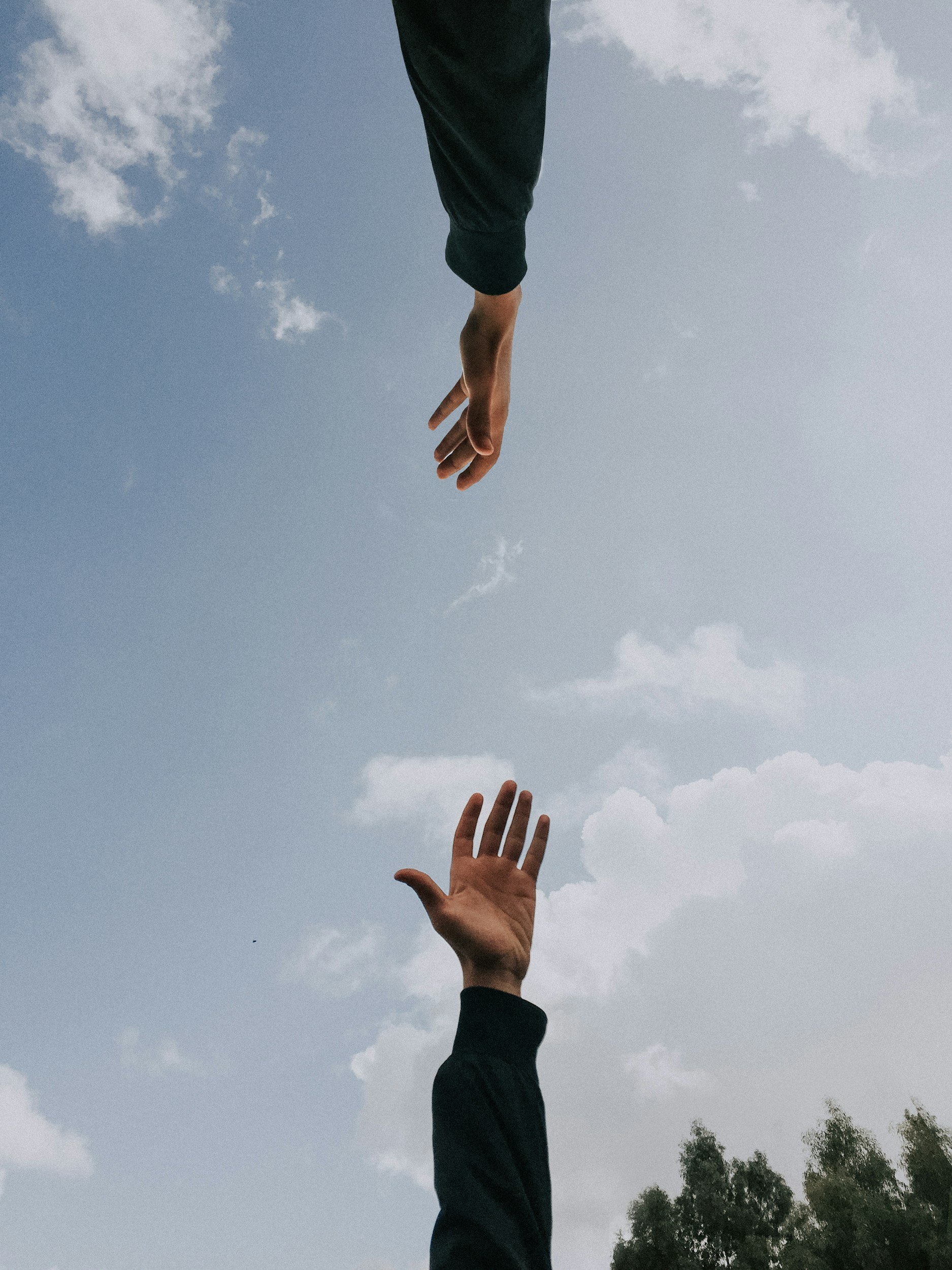 A person's hand reaches upward with fingers spread, reaching toward a sky with scattered clouds. Another hand appears in the background, also reaching upward, set against a backdrop of cloudy sky and some trees at the bottom right.