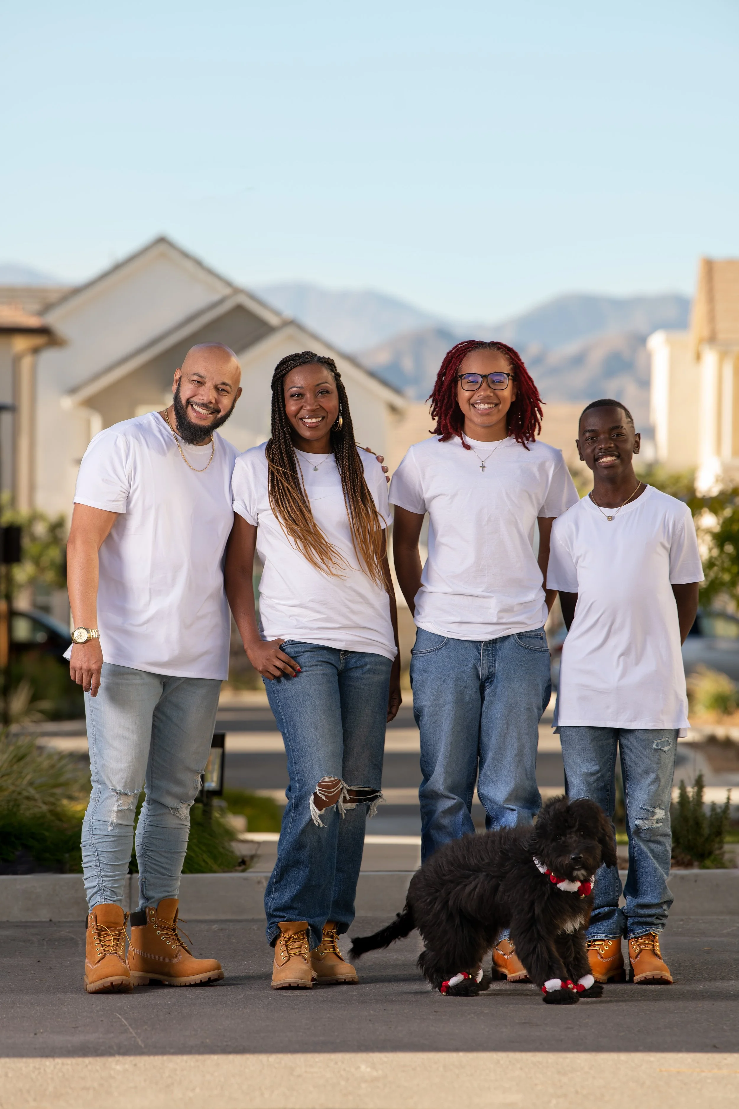 Happy family standing outdoors on a sunny day, with a mountain and a house background, and a black dog with red and white accessories in front.