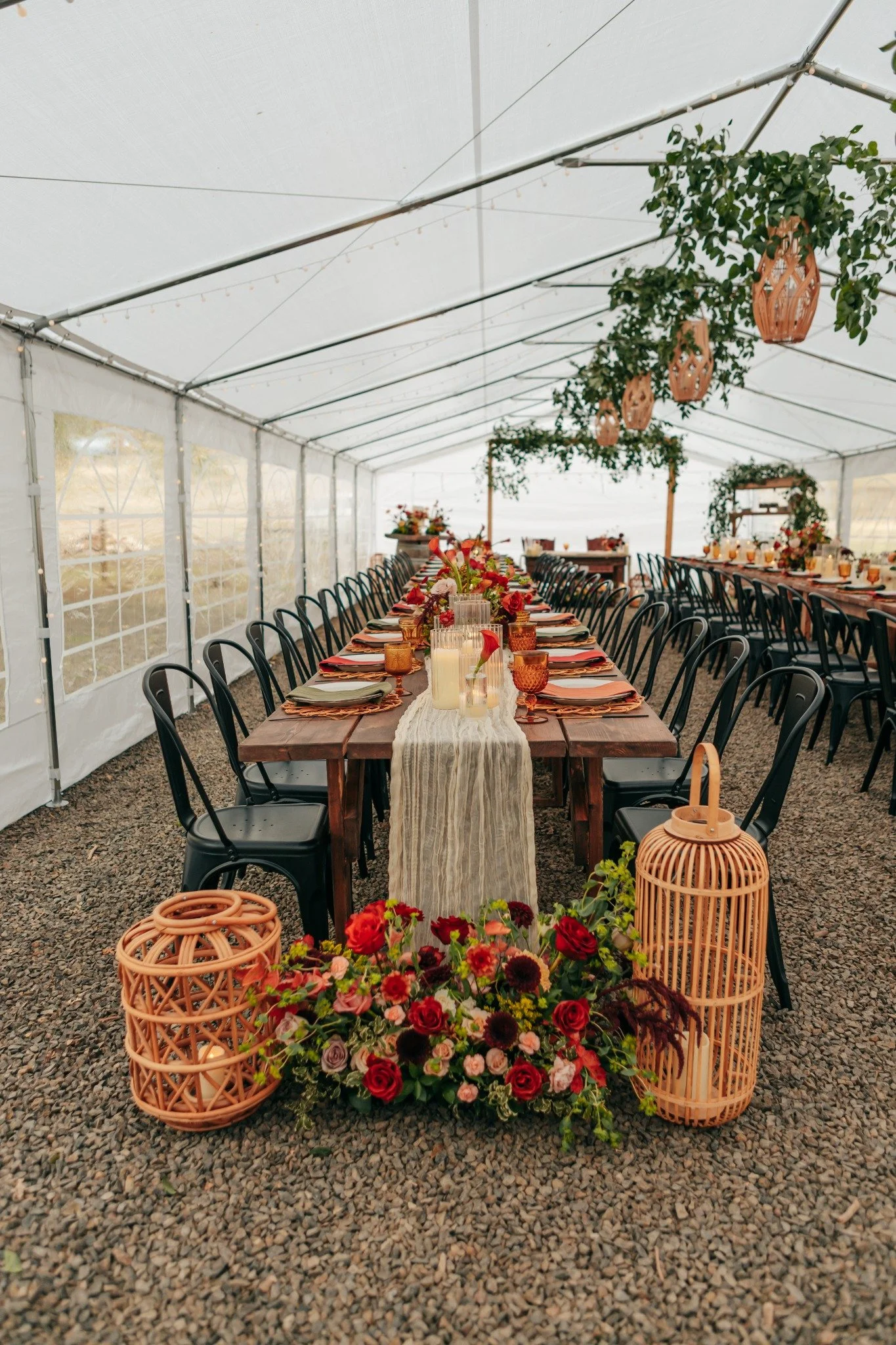 Decorated long dining table inside a white tent with hanging greenery, pink lanterns, floral centerpieces with candles, and black chairs, set for an event or celebration.