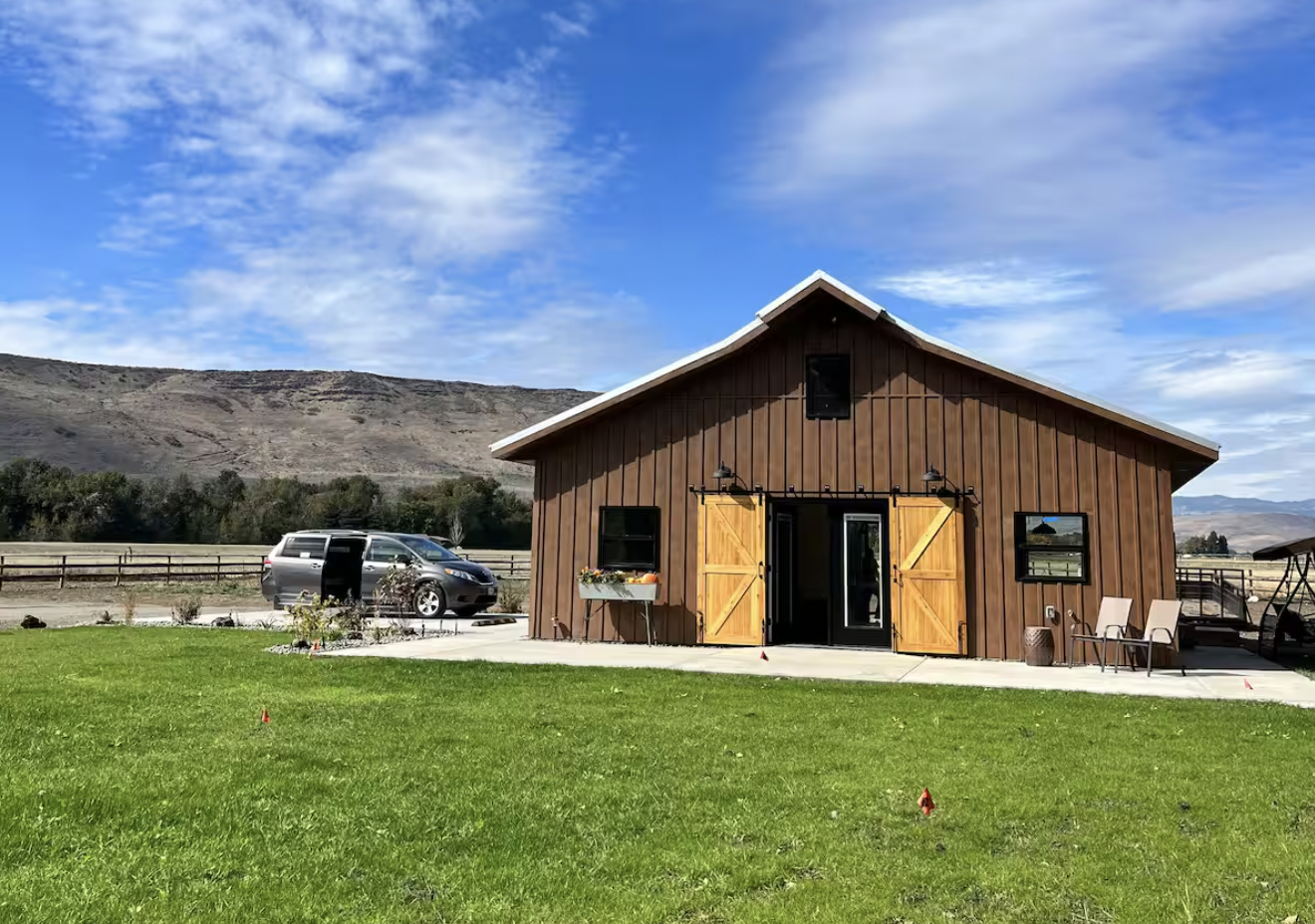 A rustic barn-style building with wooden doors and trim, sitting on a grassy lawn under a blue sky with clouds. A vehicle is parked beside the building.
