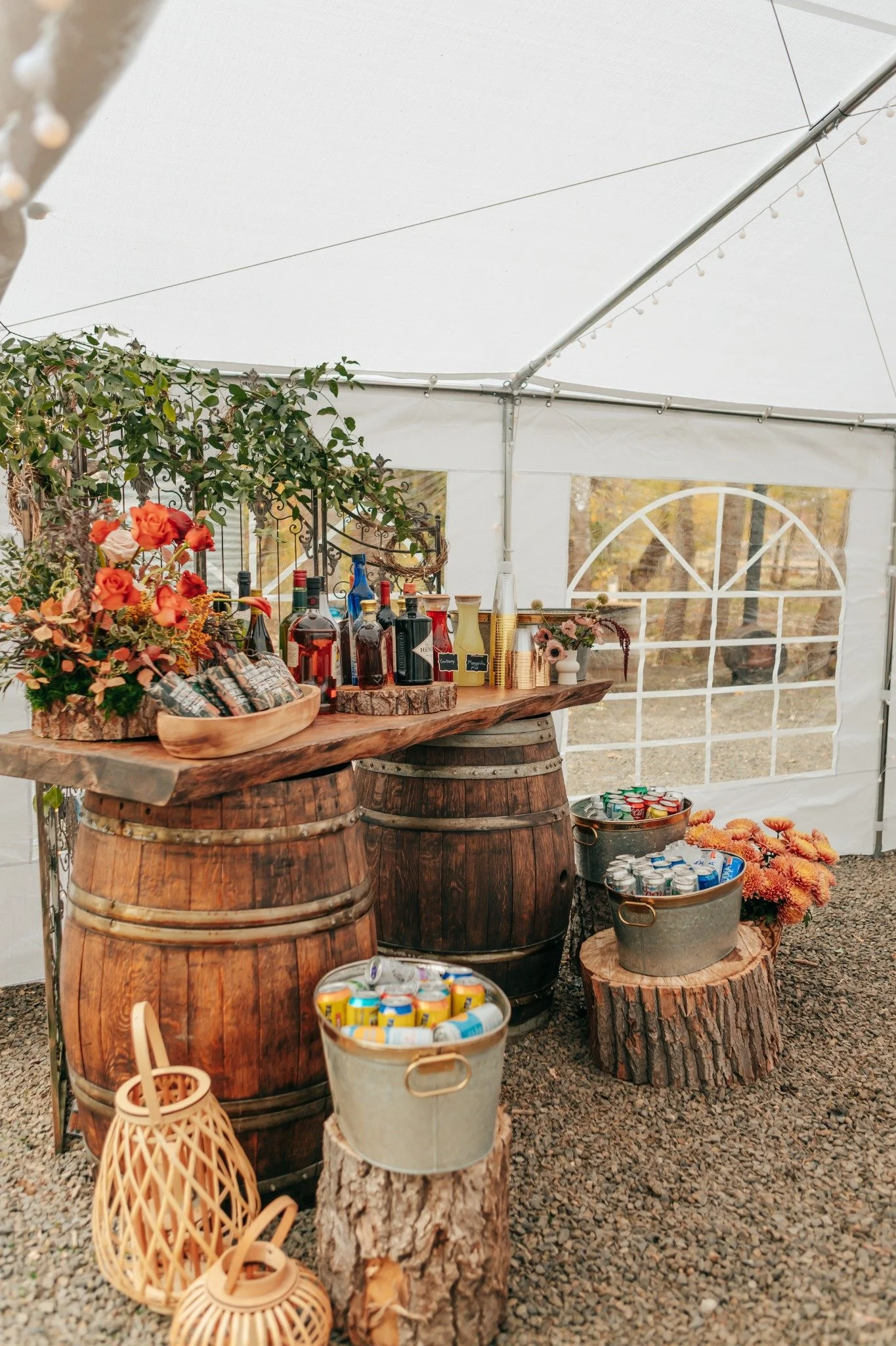 A rustic outdoor bar setup inside a white event tent, featuring wooden barrel tables, a variety of liquor bottles, canned drinks, and decorative flowers in baskets with a gravel ground.