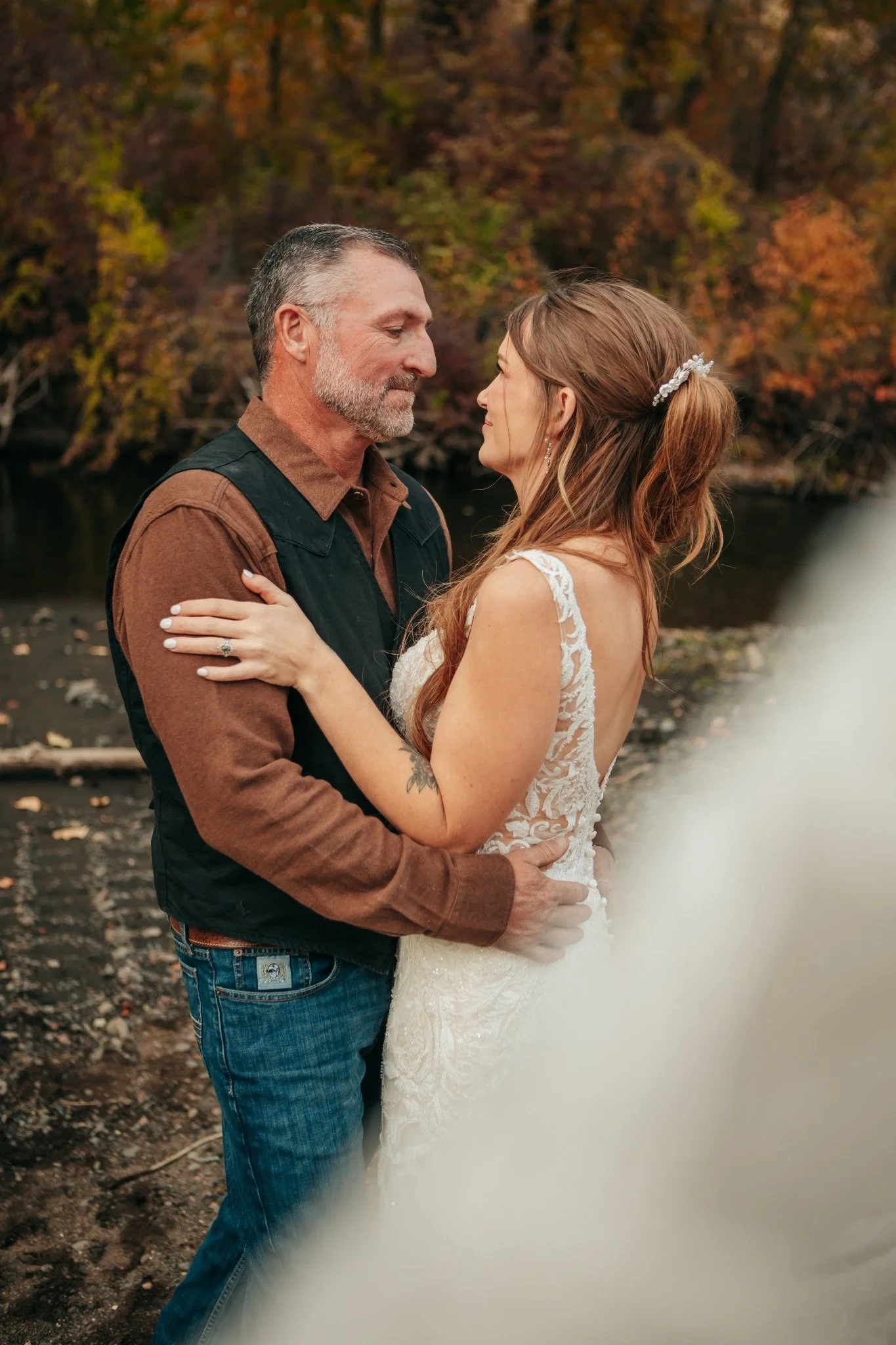 A couple dressed in wedding attire standing close together outdoors, surrounded by autumn trees and a river.