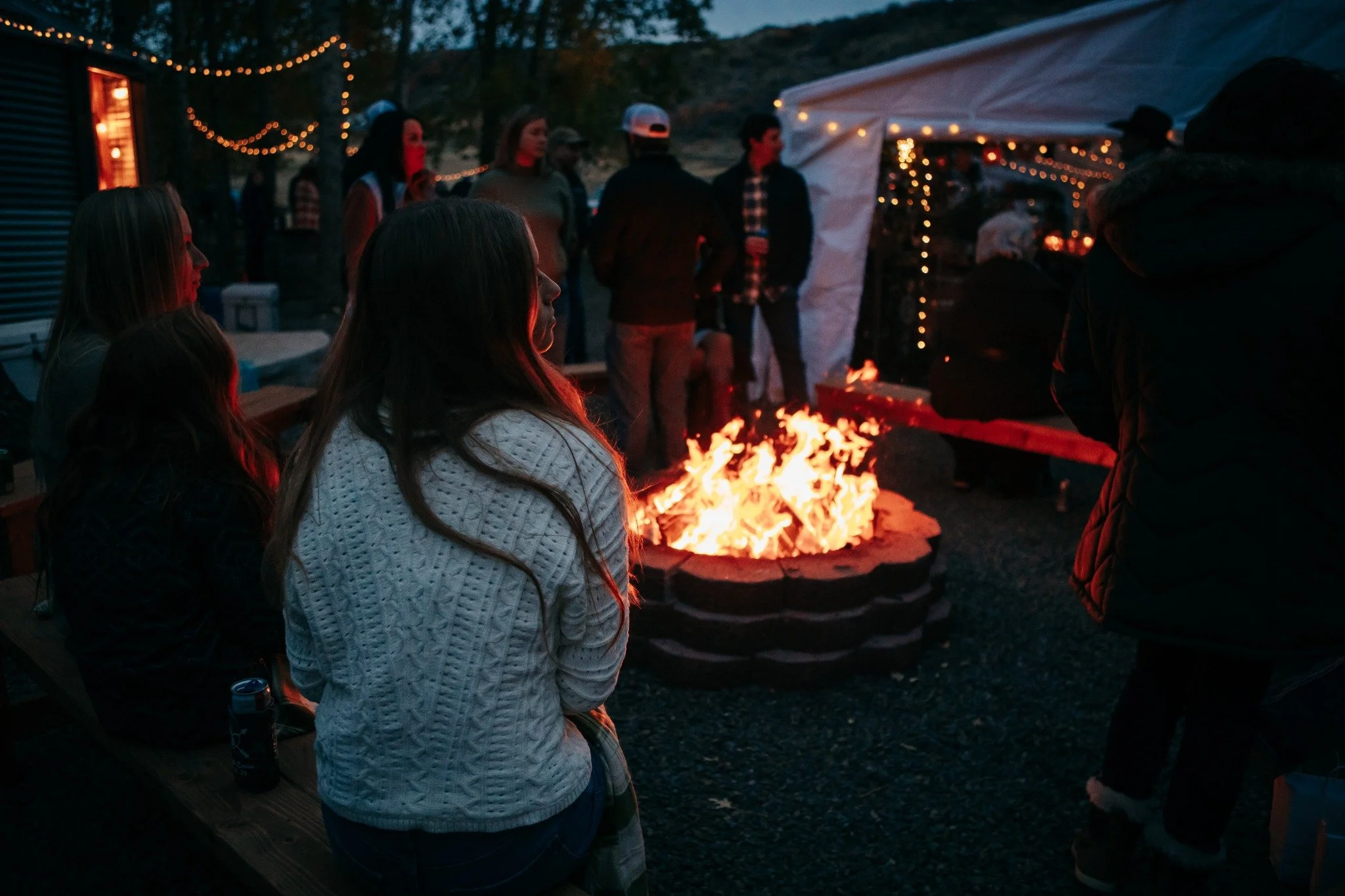 People gathered around a campfire outdoors during twilight, with string lights and a white canopy tent in the background.