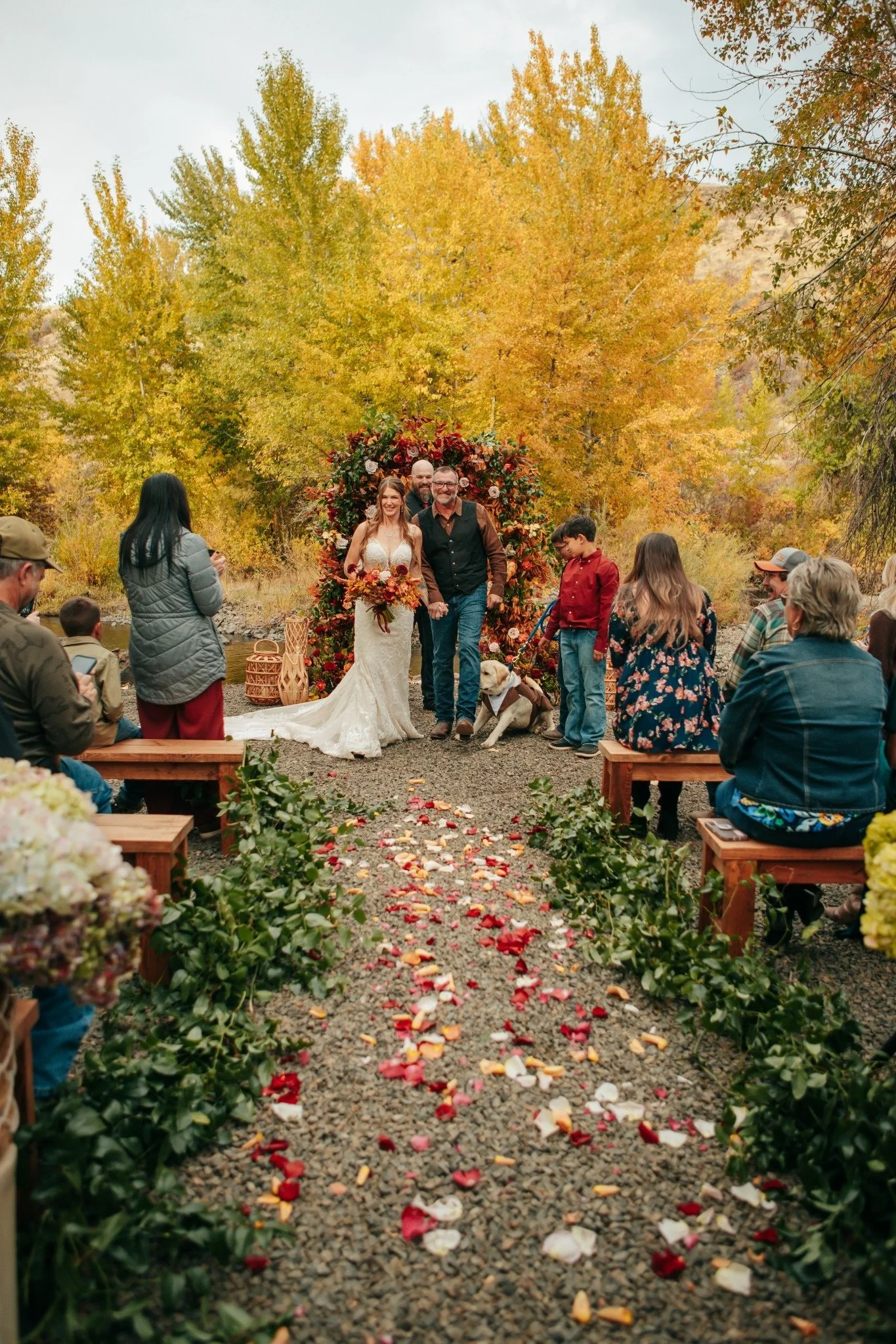 A wedding ceremony outdoors during autumn with trees in yellow and orange leaves. The bride in a white wedding dress and the groom holding hands are walking down an aisle decorated with rose petals, alongside a man, and surrounded by guests sitting o
