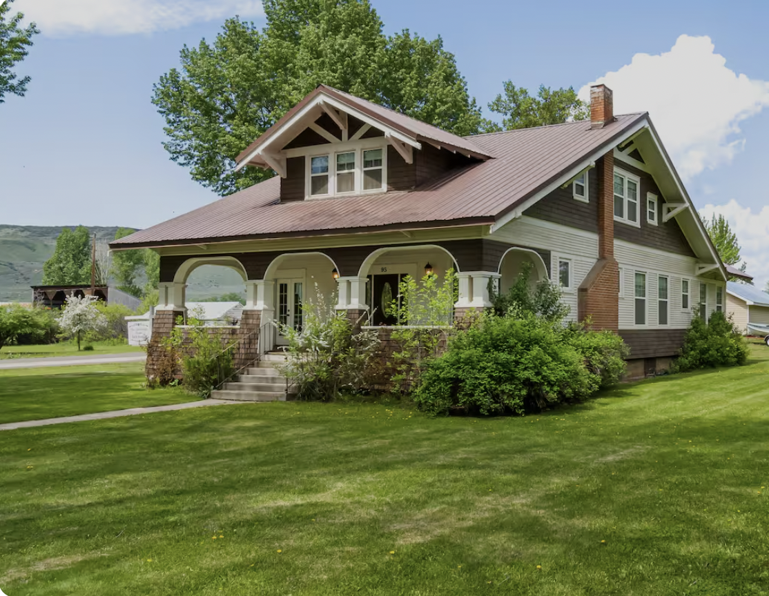 A large two-story house with a brown roof, white and dark wood siding, and a front porch with stone columns, surrounded by green lawn and trees.
