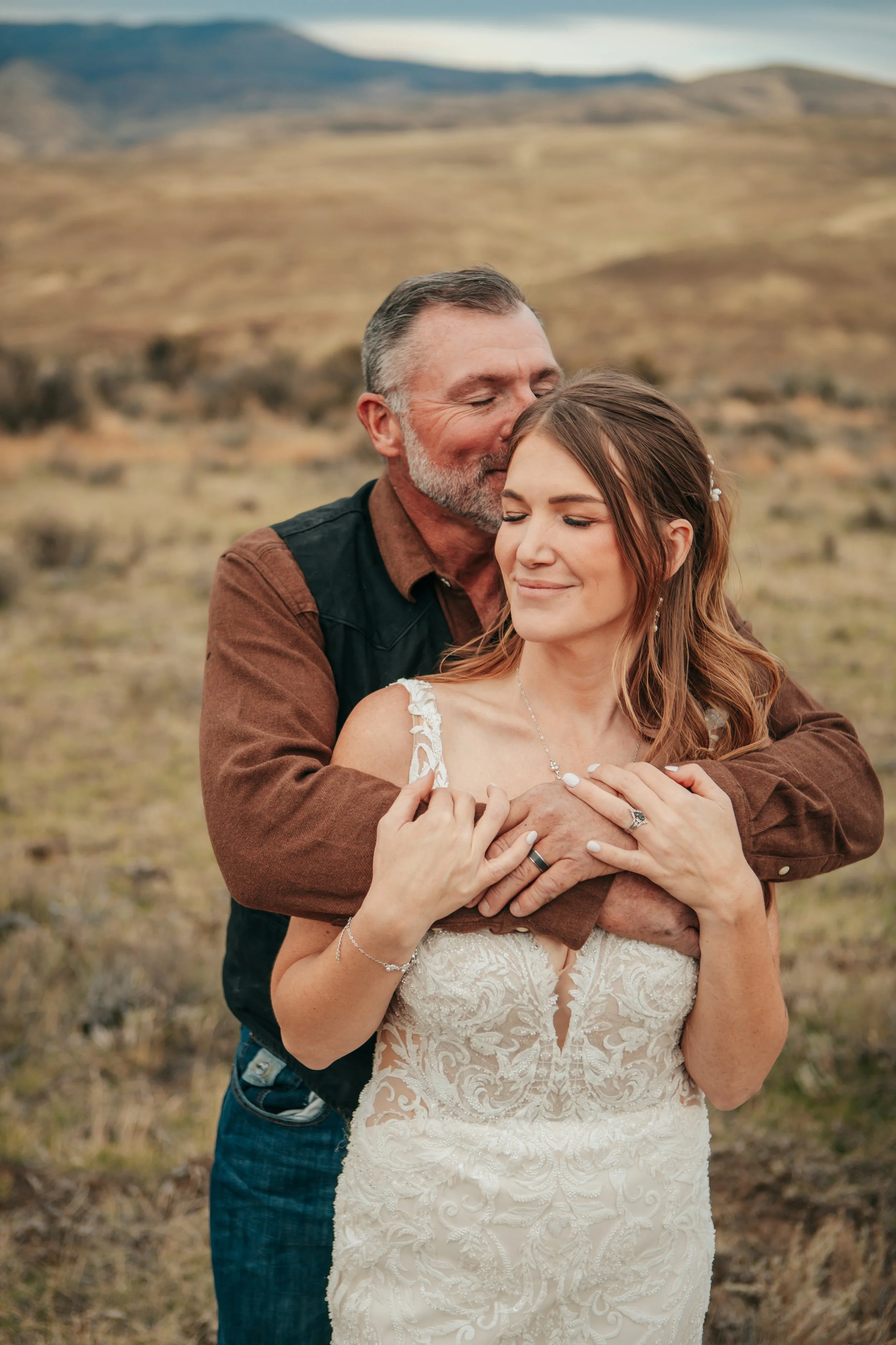 A couple hugging outdoors with a scenic mountainous landscape in the background. The man is kissing the woman's forehead with his eyes closed, while she has a gentle smile and her eyes closed. The woman is wearing a lace wedding dress, and the man is dressed casually in a brown shirt with a black vest.