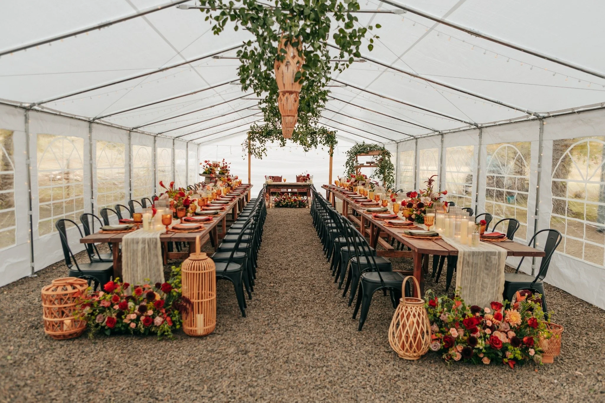Inside of a decorated wedding or event tent with long wooden tables set with floral arrangements, candles, and tableware. Greenery and floral decor hang from the ceiling, and baskets with flowers are placed at the entrance. The tent has clear plastic