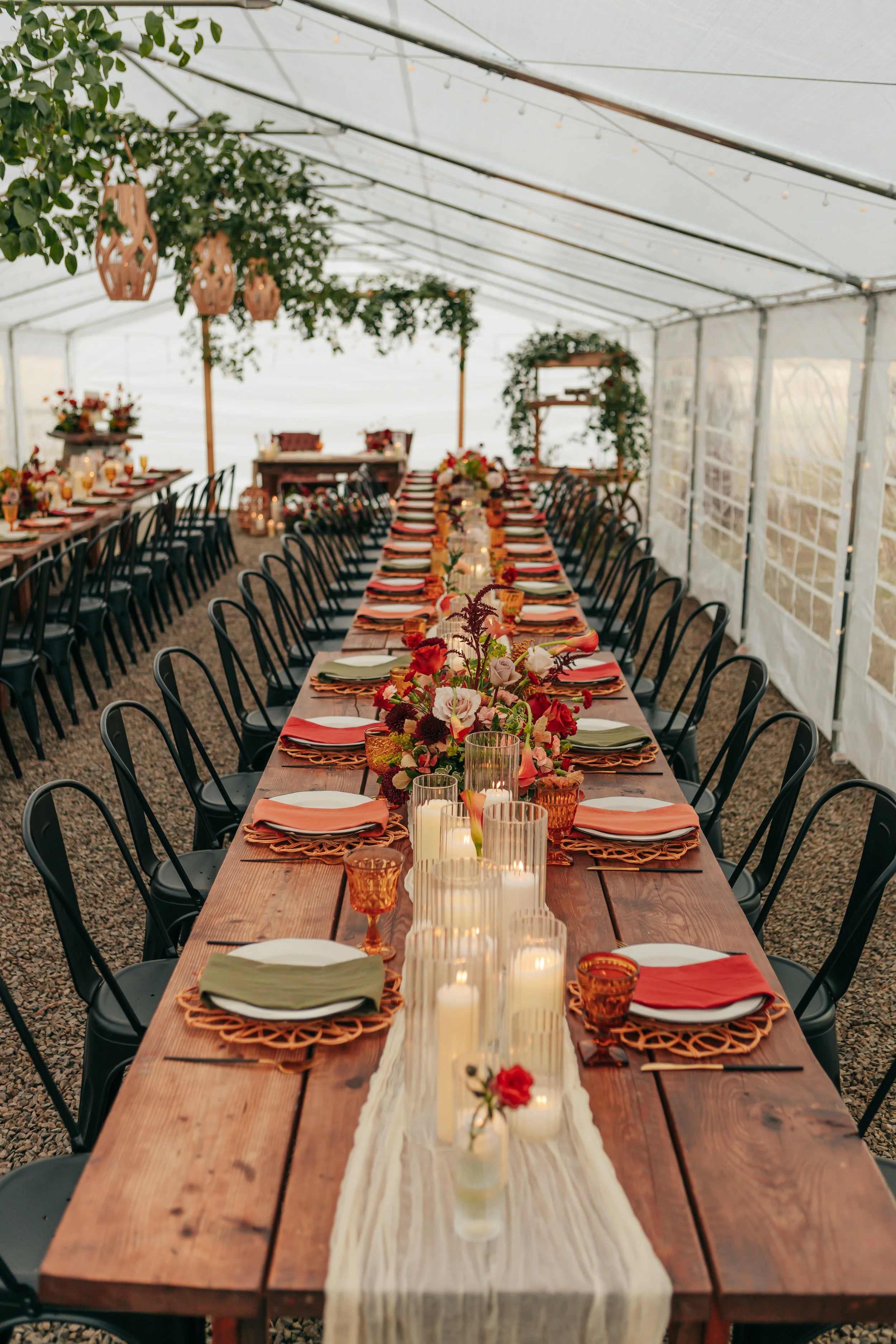 A long rustic wooden table set for a formal event in a covered outdoor space with string lights, floral centerpieces, candles, and colorful place settings.