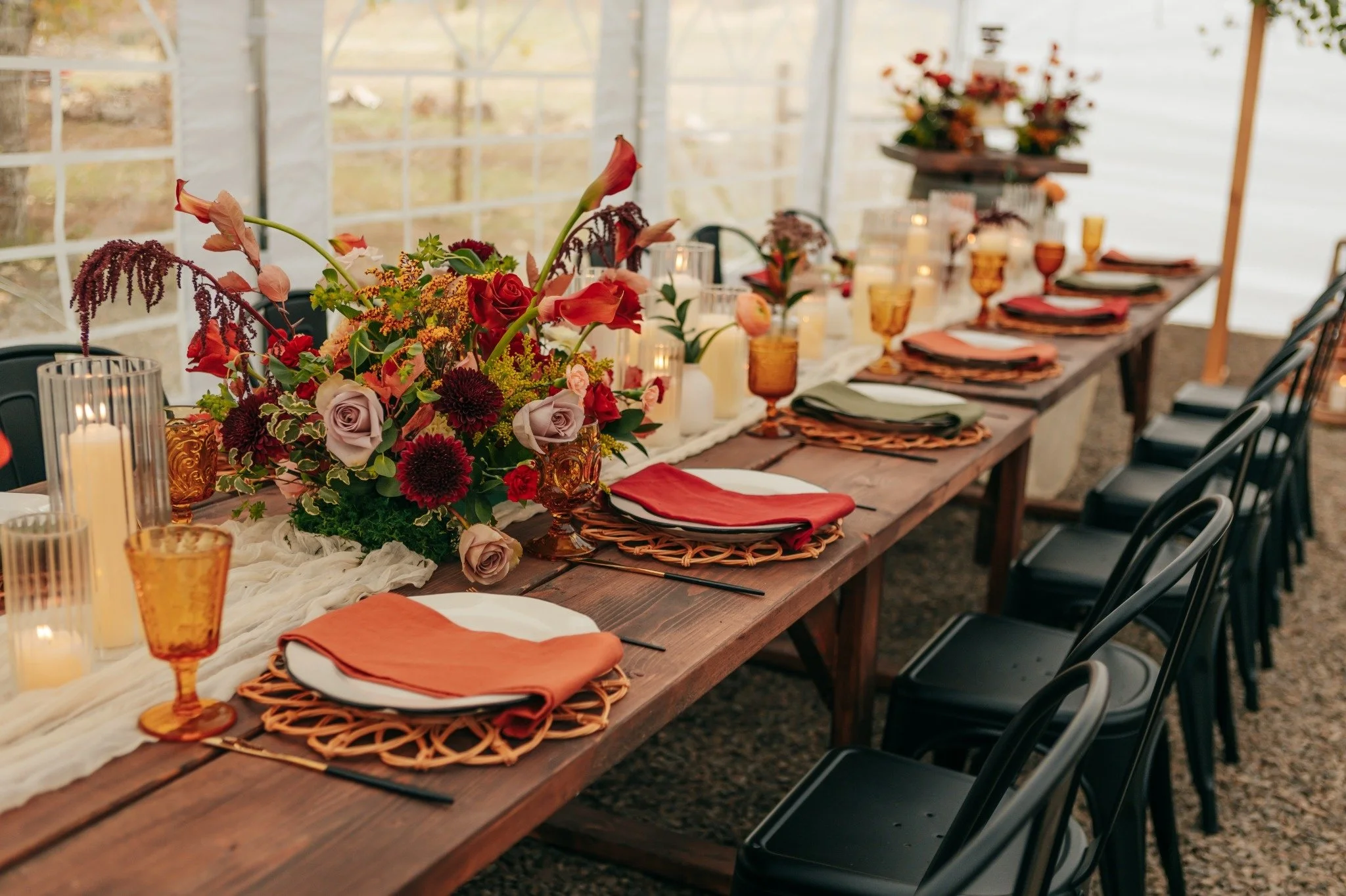 A long wooden banquet table decorated with a large vibrant floral centerpiece with roses, calla lilies, and other flowers, surrounded by wine glasses, candles, and folded napkins in warm colors, set for a gathering inside a bright greenhouse or conservatory with large windows.