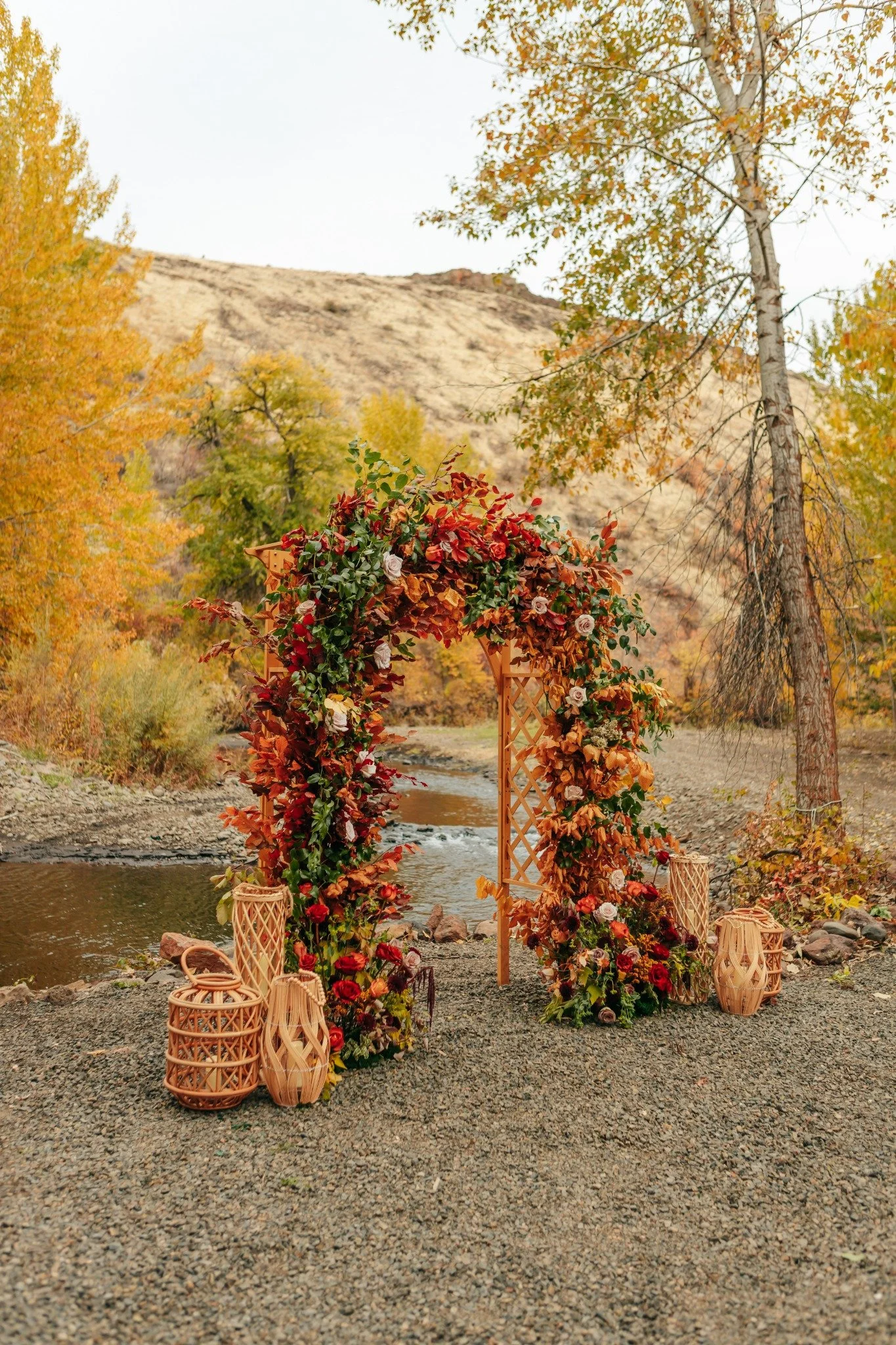 Outdoor wedding arch decorated with flowers and leaves, set next to a river with autumn trees and a mountain in the background.