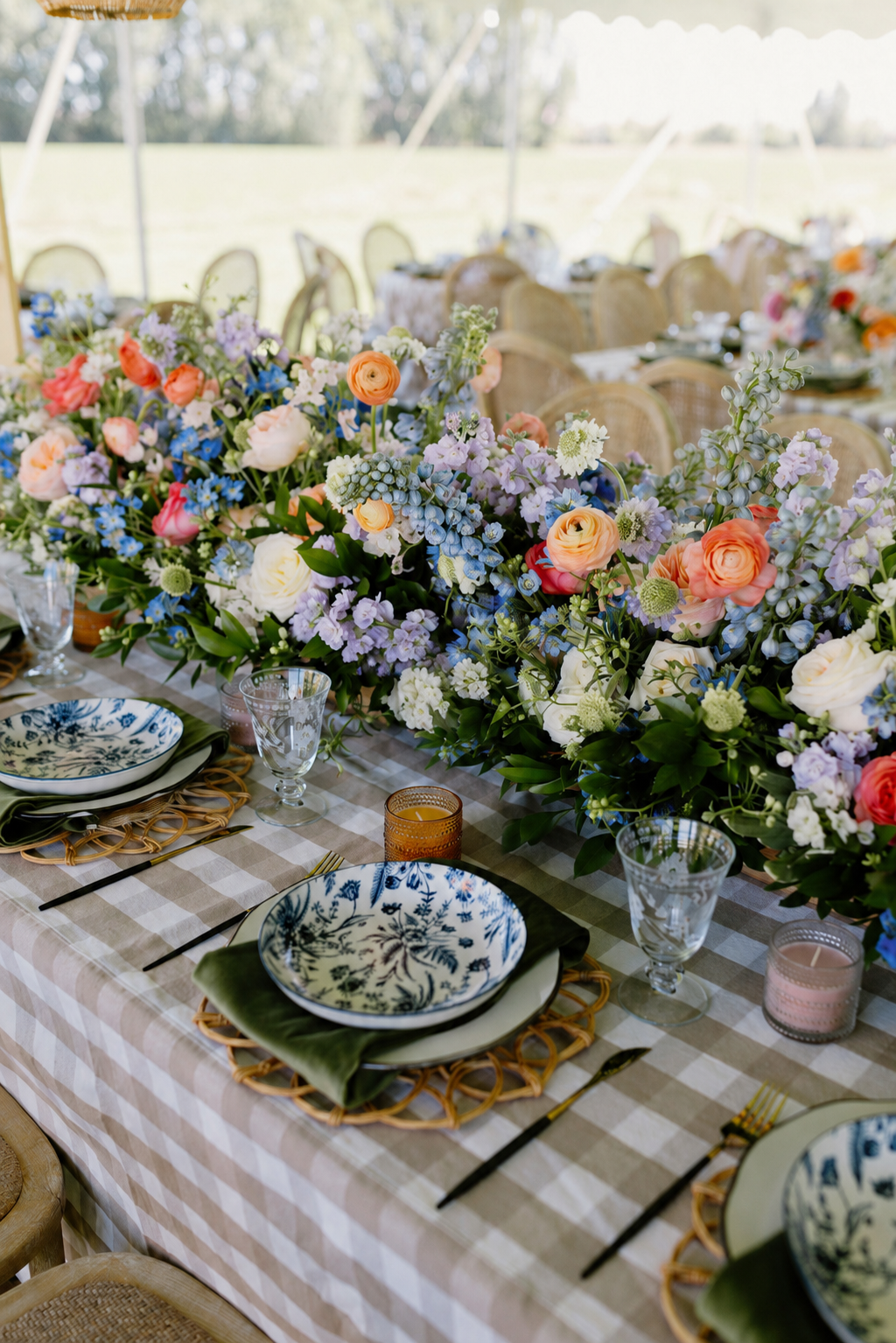 Table decorated with a large floral centerpiece of mixed colorful flowers, set with blue-and-white patterned plates, gold flatware, glassware, and small candles in amber and pink holders, in an outdoor setting.