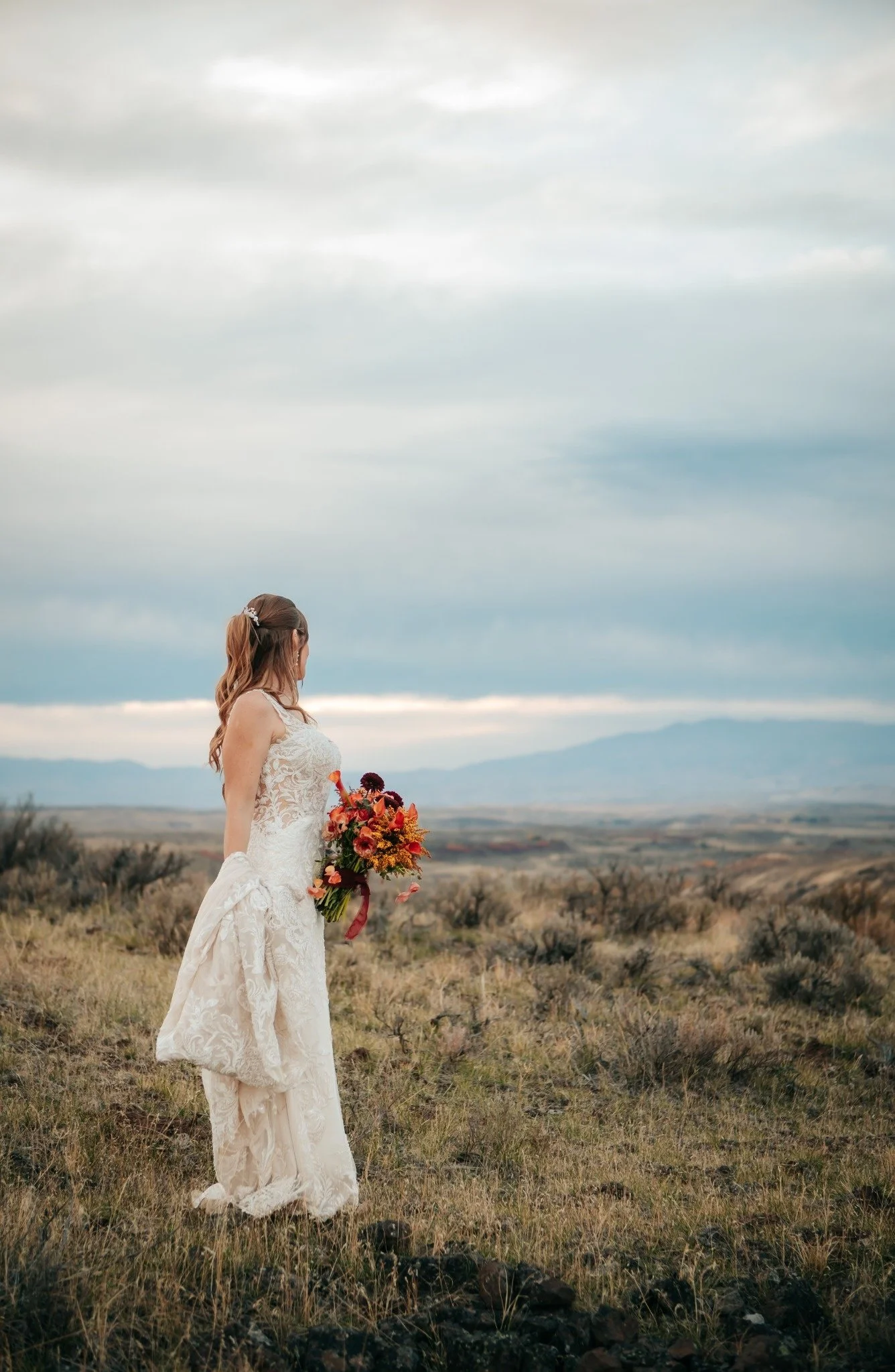 A woman in a lace wedding dress holding a large colorful bouquet stands in a vast open field with mountains and cloudy sky in the background.