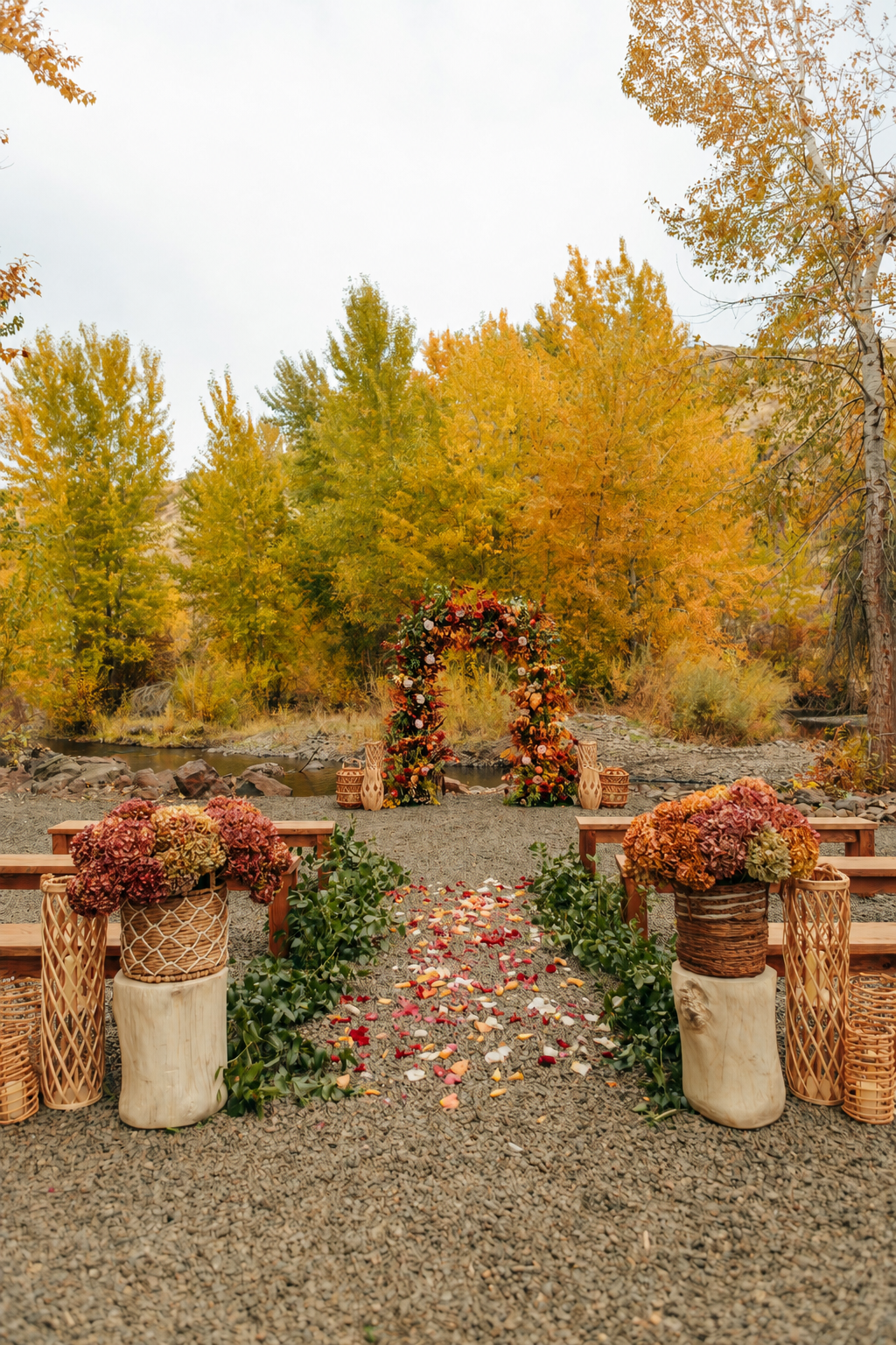 Outdoor wedding setup with a floral arch over a gravel pathway, surrounded by autumn trees, flower arrangements, candles, and benches.