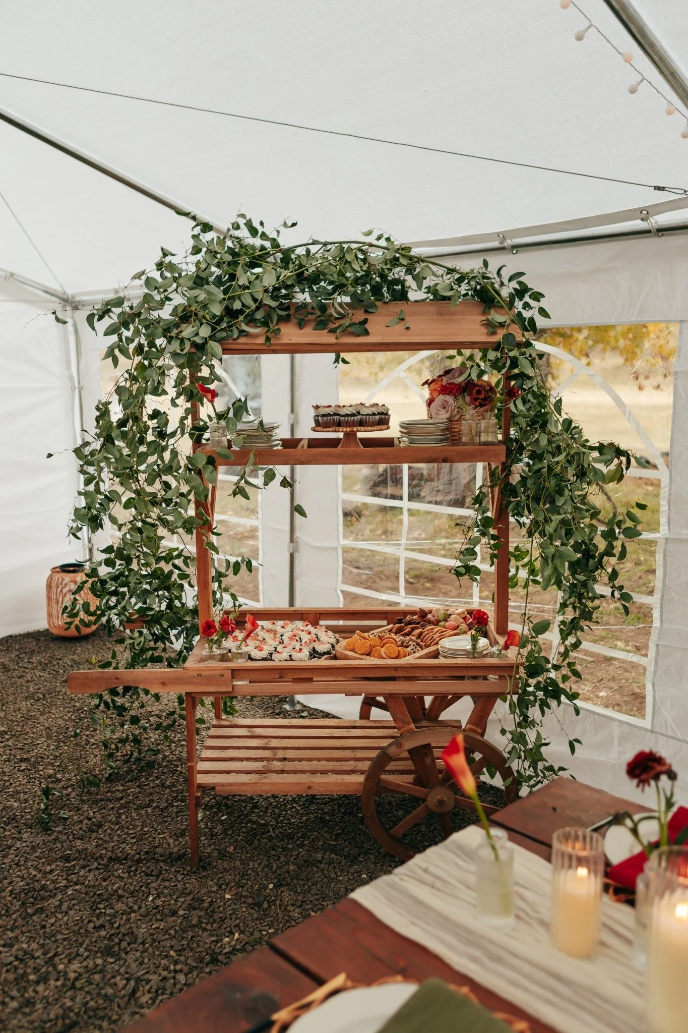A decorated food table inside a white tent, featuring a display of cupcakes, cookies, and pastries, surrounded by greenery and floral arrangements, with candles and small decor items.