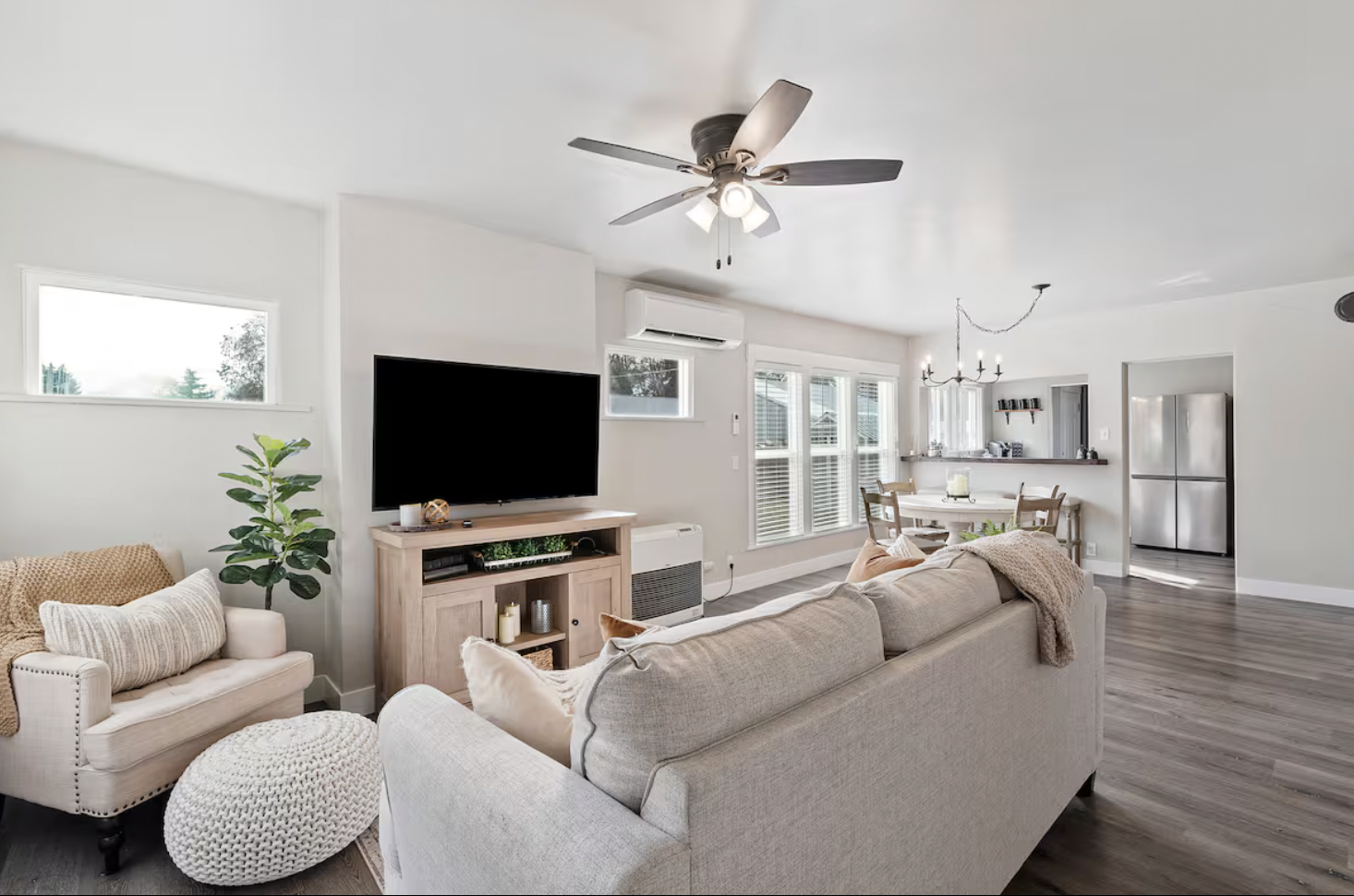 Bright living room with beige sofa, armchair, large flat-screen TV, wooden TV stand, green potted plant, windows, ceiling fan, dining table with chairs, and open kitchen with stainless steel refrigerator.