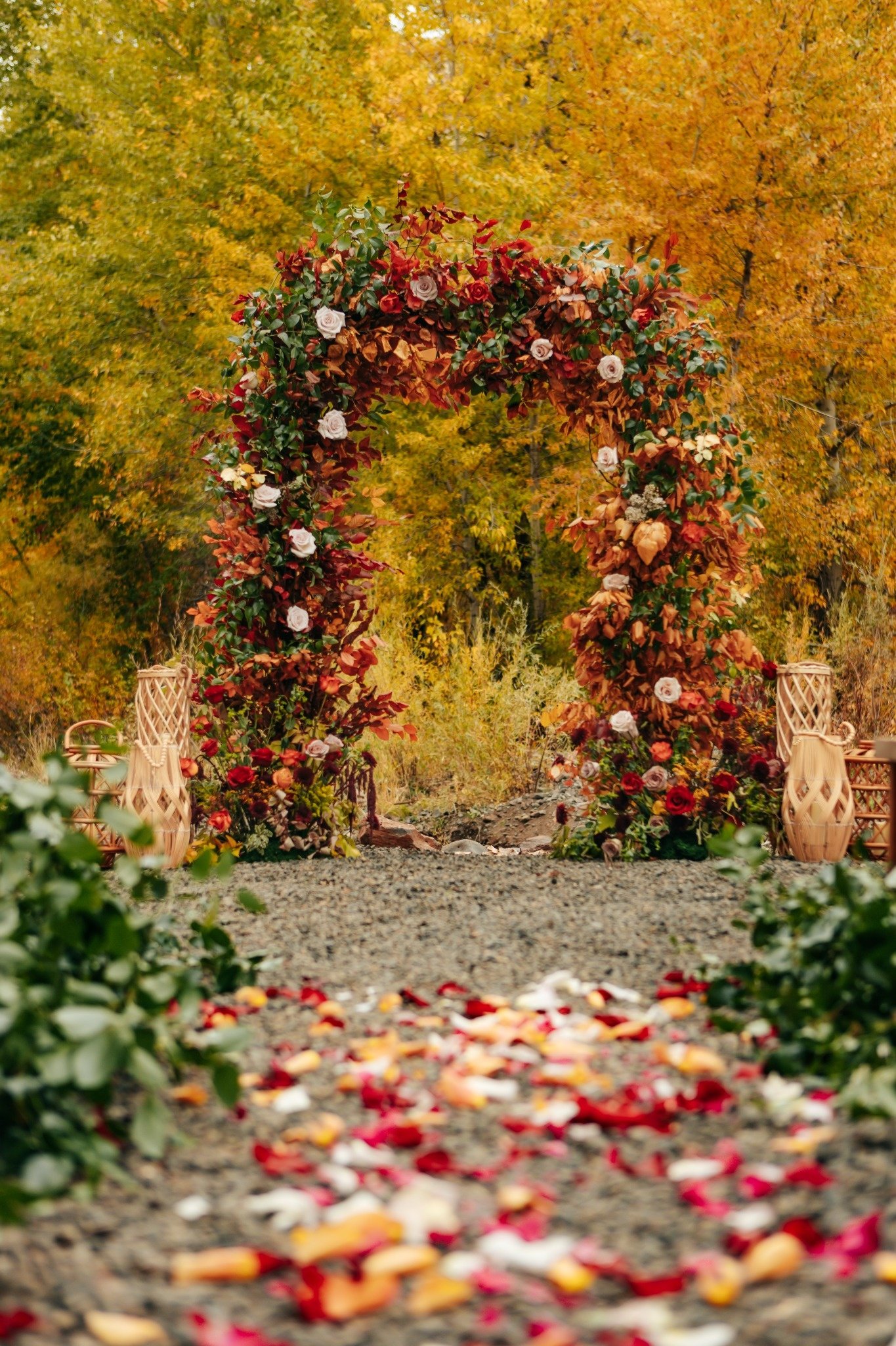 Autumn outdoor ceremony setup with a flower-covered arch, surrounded by fallen flower petals and chairs, against a backdrop of colorful fall trees.