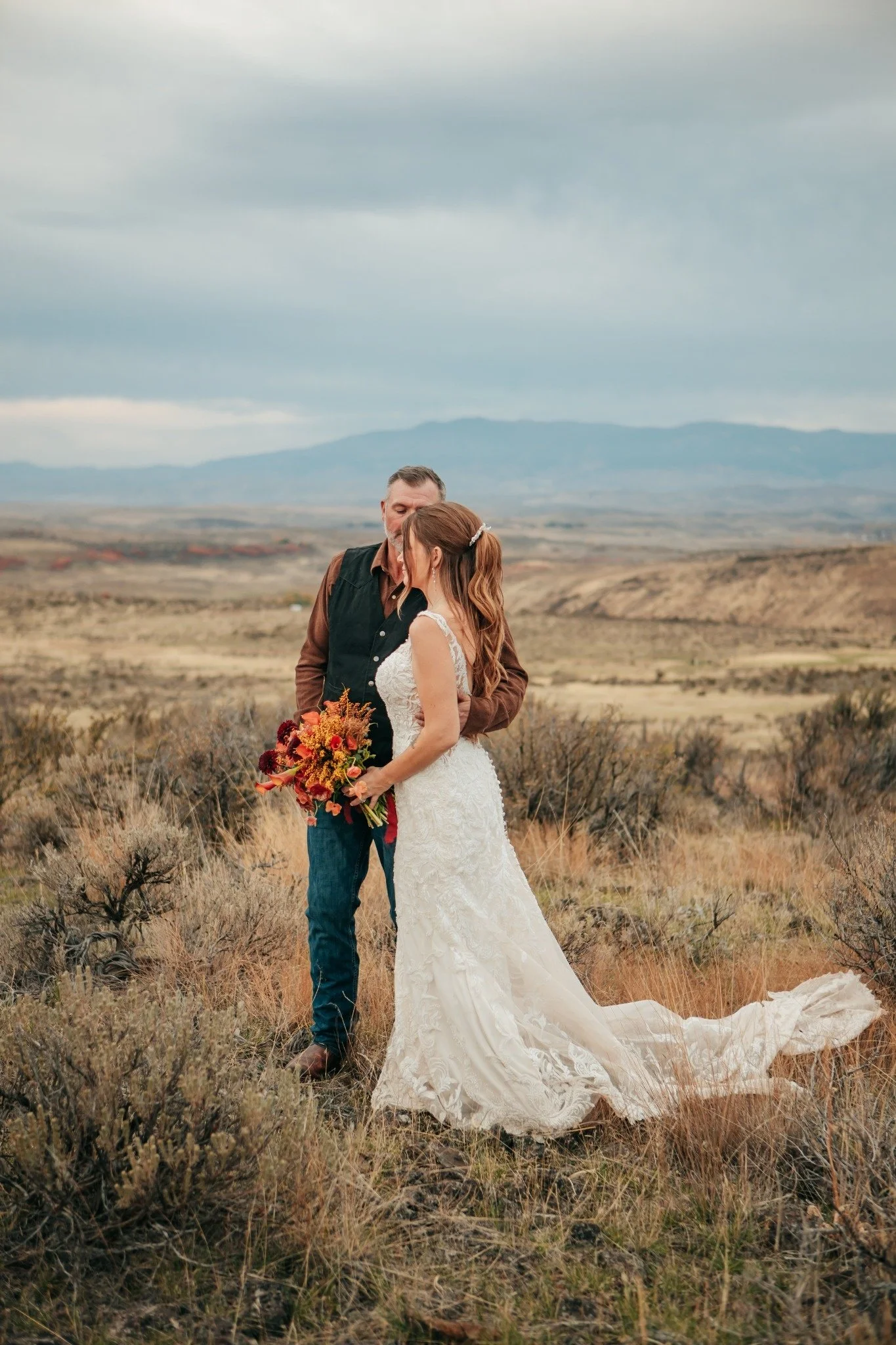 A bride and groom standing close together in a field with dry grass and shrubs, embracing each other. The bride is holding a bouquet of flowers and is wearing a white lace wedding dress. The groom is wearing a brown shirt with a black vest and blue j