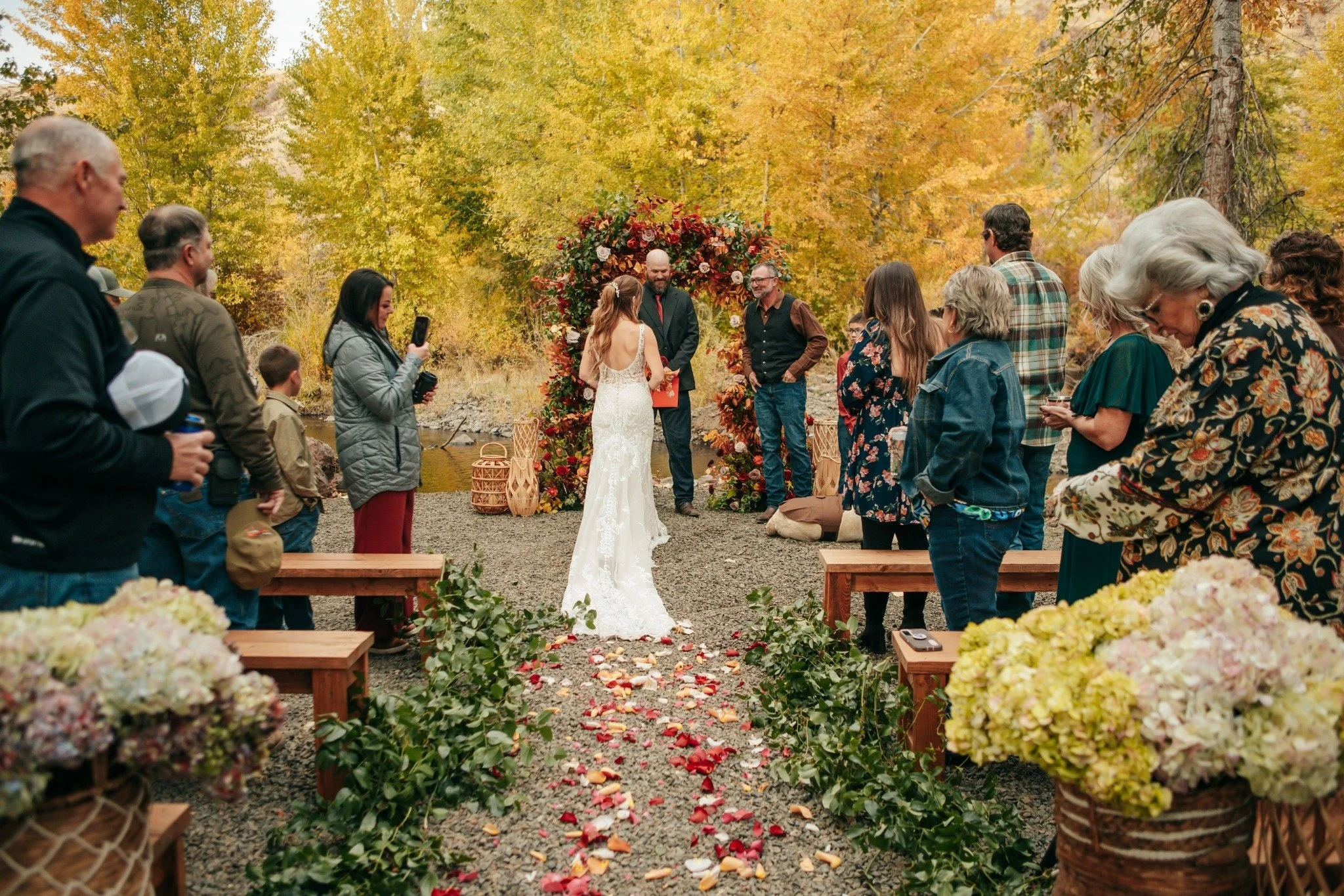 A wedding ceremony outdoors with a bride and groom standing under a floral arch surrounded by guests, set against fall foliage.