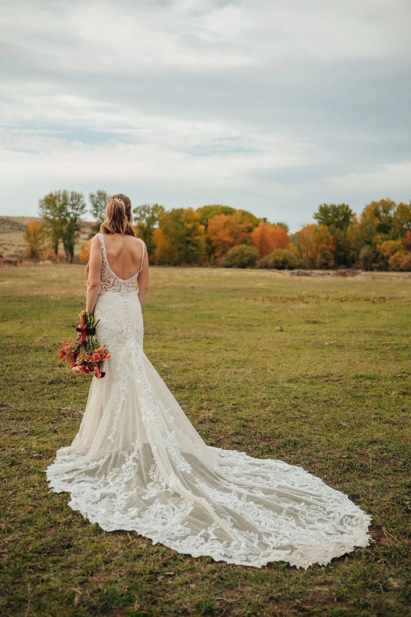 A woman in a wedding dress standing outdoors on a grassy field, holding a bouquet, with trees displaying fall foliage and a cloudy sky in the background.