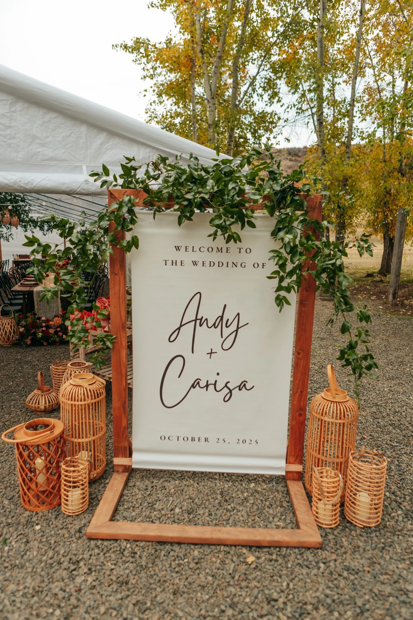 A wedding welcome sign with green foliage hanging on a wooden frame, set outdoors with fall trees in the background. The sign reads: "Welcome to the wedding of Andy + Carisa, October 25, 2025." Surrounding the sign are woven lanterns on a gravel surf
