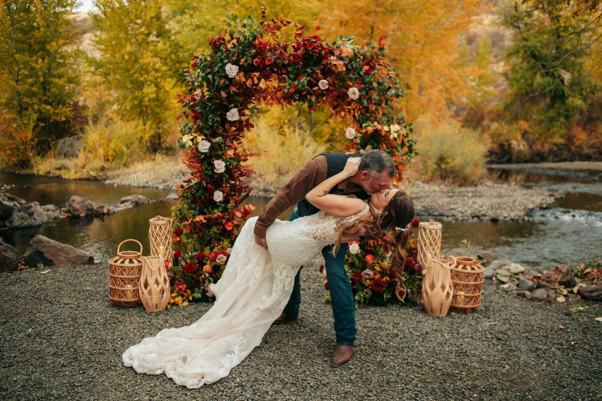A couple kissing during their outdoor wedding ceremony by a river, with autumn-colored trees in the background and a decorated floral arch.