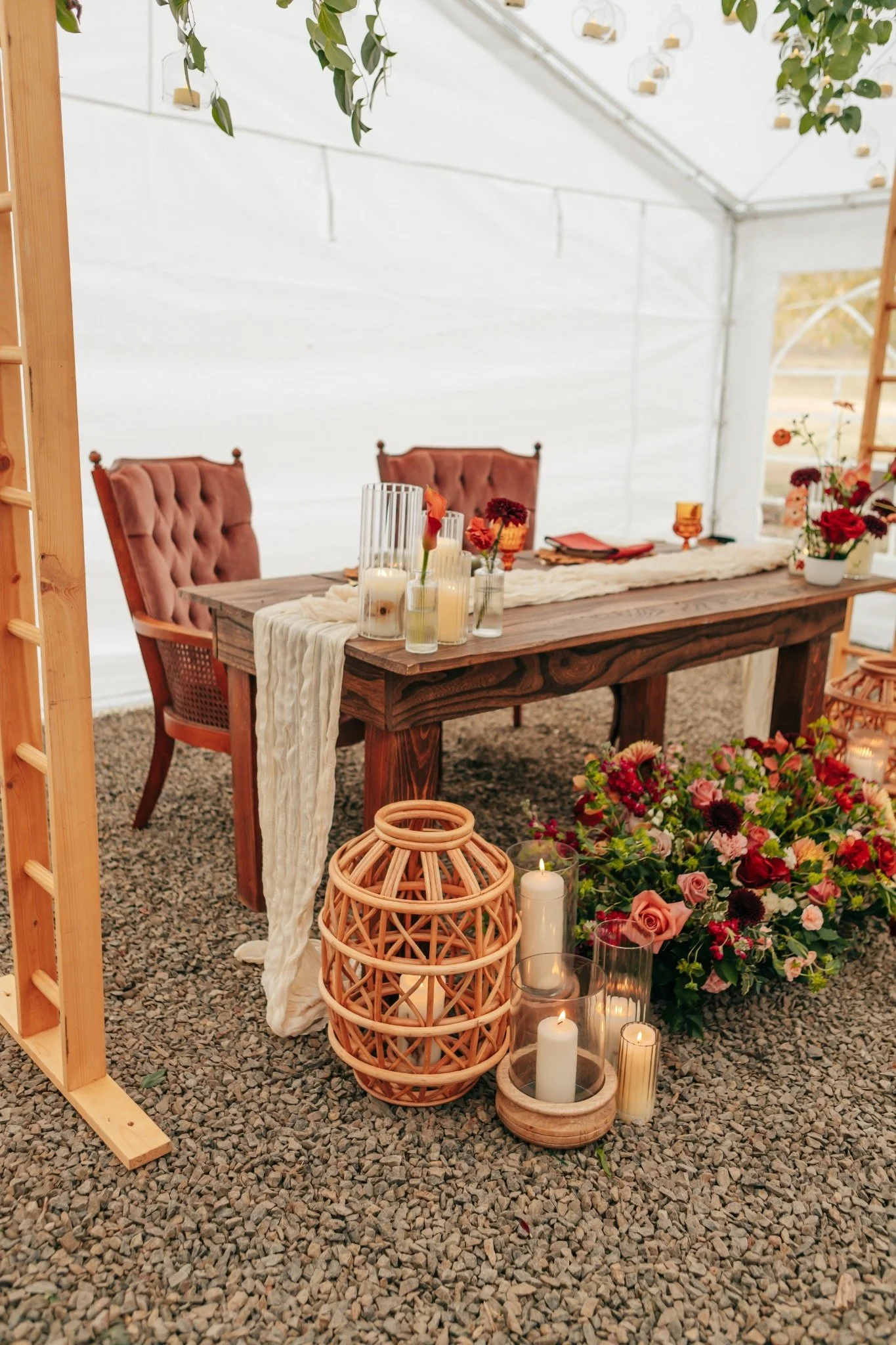 A rustic wooden table decorated with candles and flowers, set inside a white tent with hanging greenery and chairs in the background, creating an elegant event setting.