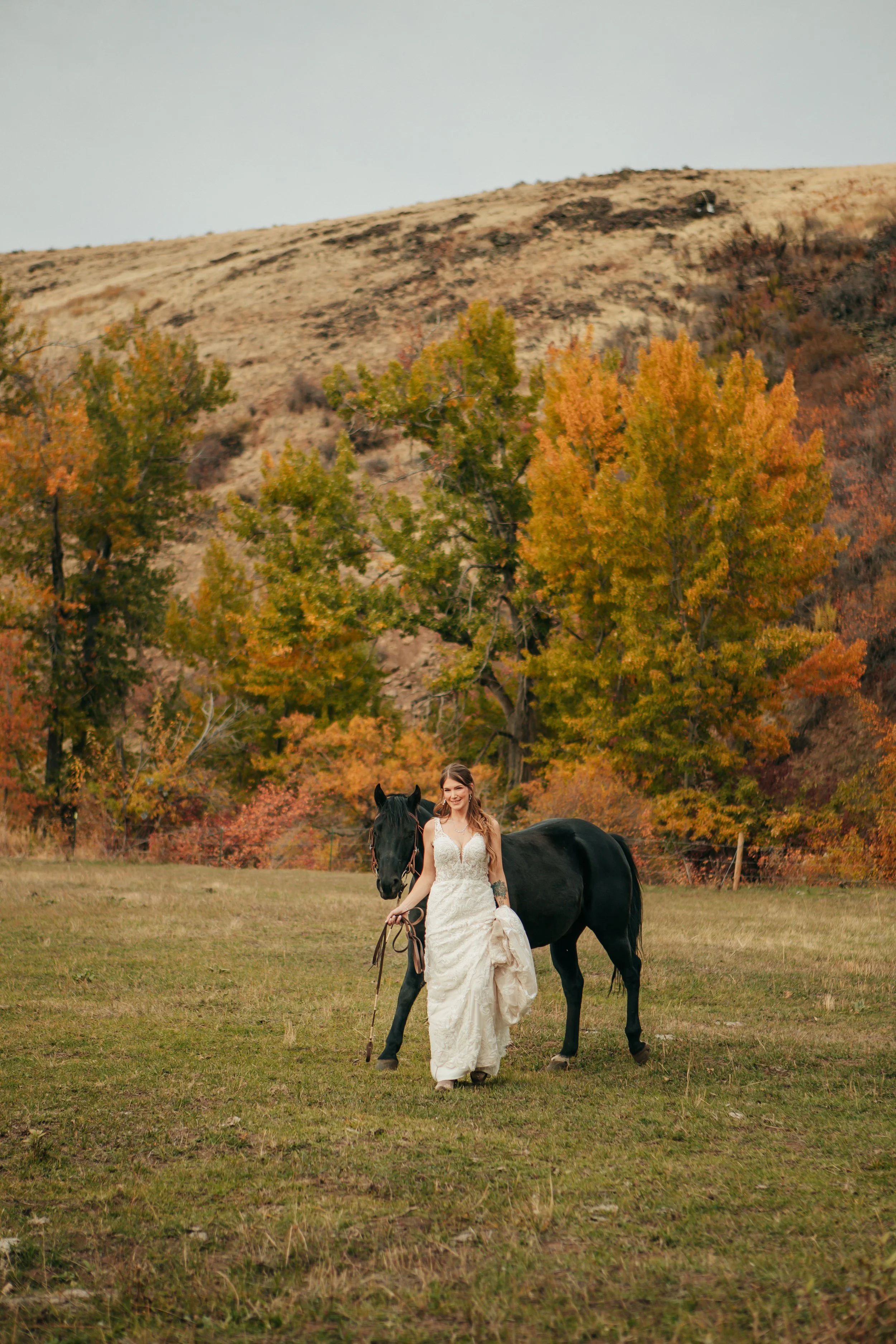 A woman in a wedding dress at Riverwild Weddings & Events standing next to a black horse with a scenic background of trees at with fall foliage and a hill.